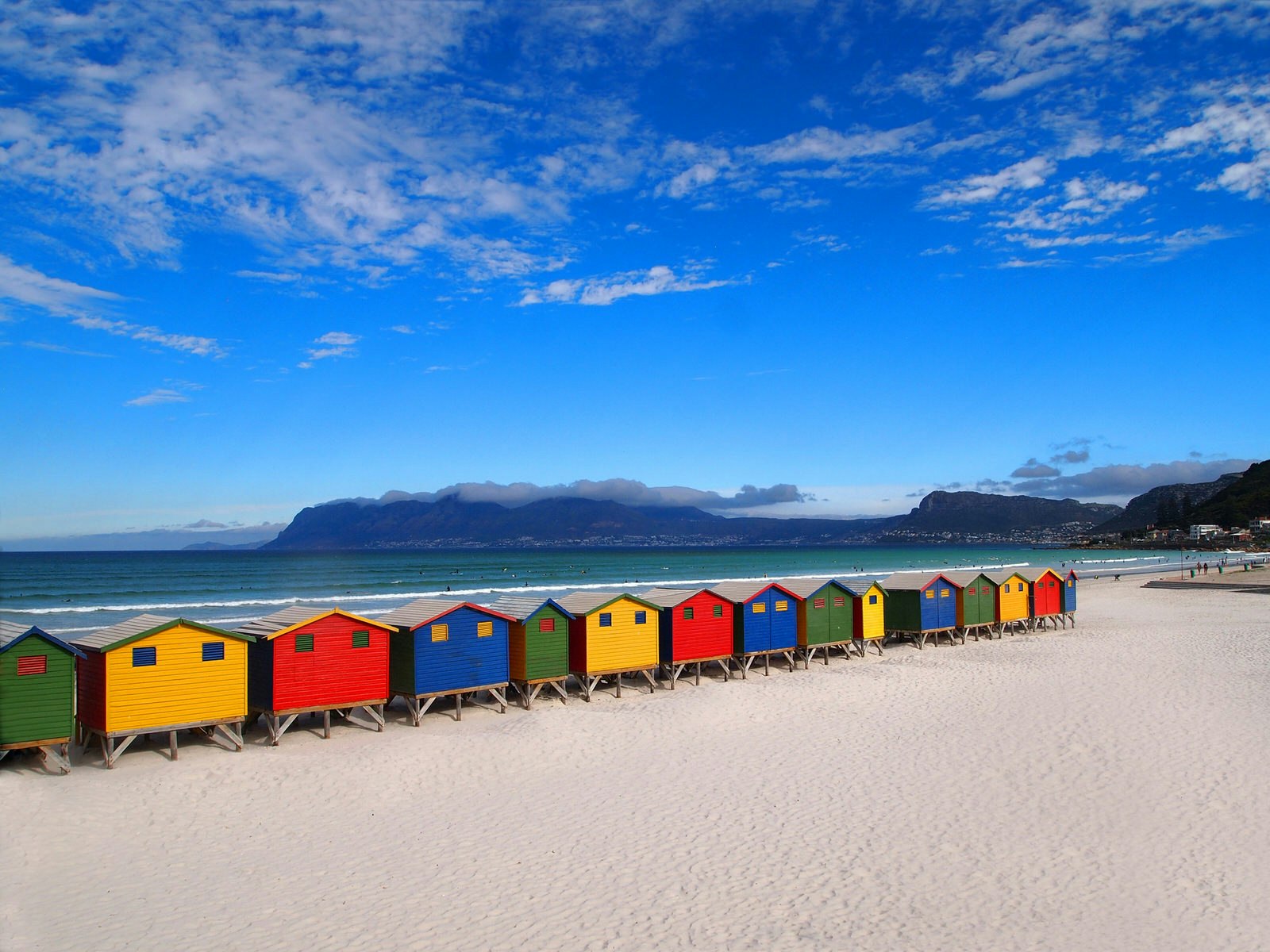 Under a deep blue sky and sitting atop a white sand beach is a line of closely-spaced beach huts. The colours of each is different, some bright yellow, others red, blue or green © Ariadna22822 / Shutterstock