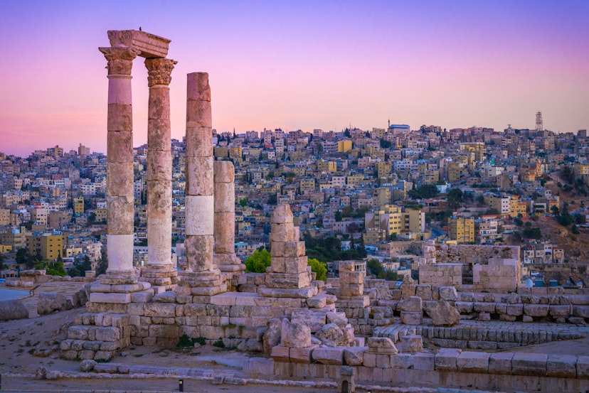 Several Roman columns stand tall and alone atop a hill, with the city in the distance as a backdrop; the sky is an incredible shad of pink and purple
