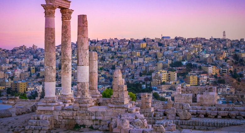 Several Roman columns stand tall and alone atop a hill, with the city in the distance as a backdrop; the sky is an incredible shad of pink and purple