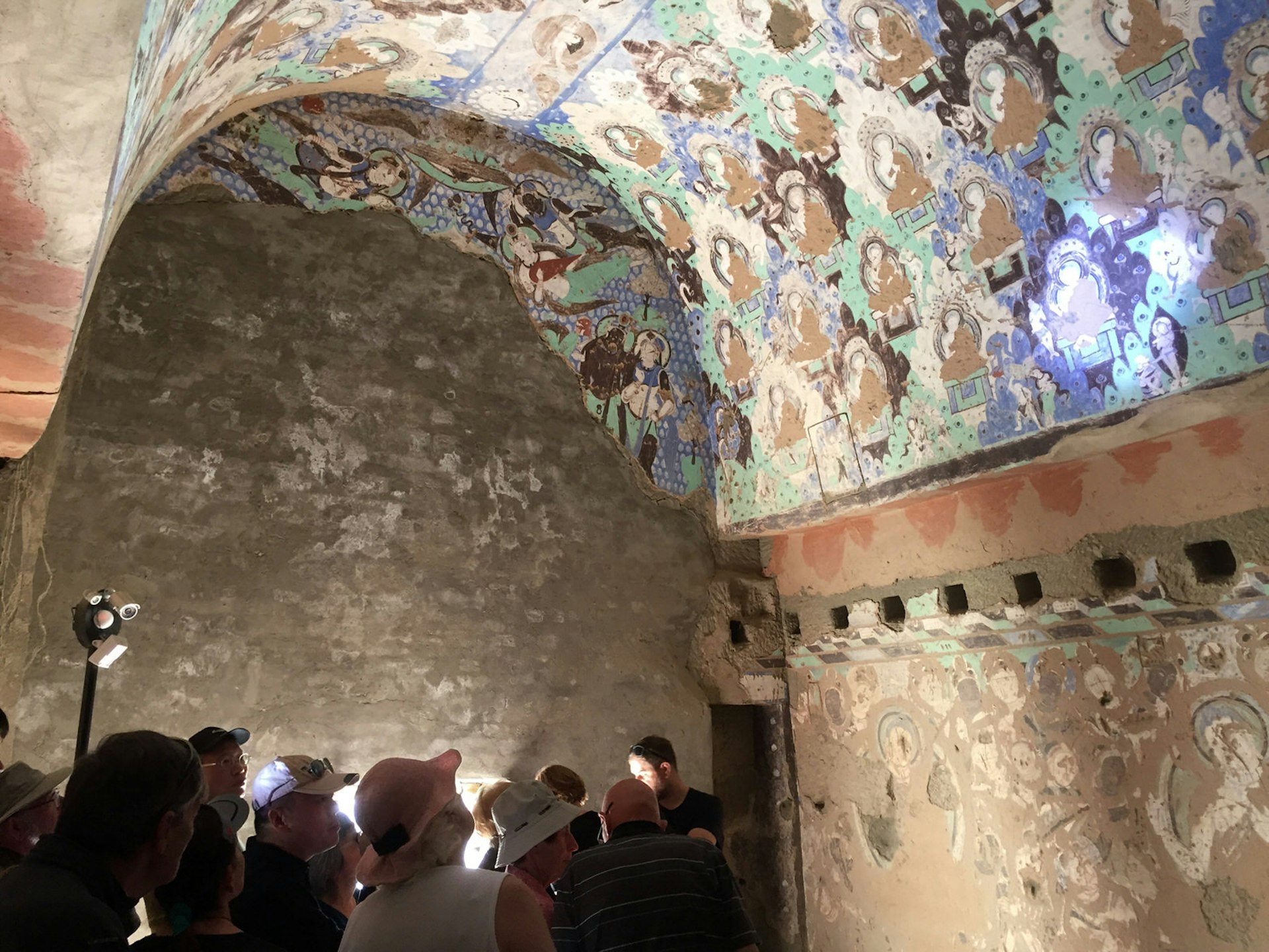 A group of visitors huddled below left stare up at an arched cave ceiling with blue, green and brown Buddhist murals illuminated by spotlights.