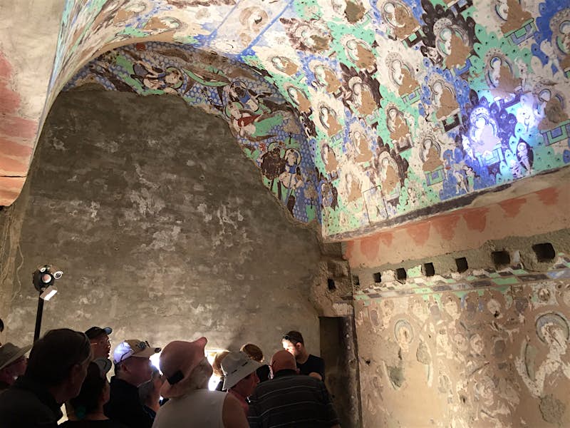 A group of visitors huddled below left stare up at an arched cave ceiling with blue, green and brown Buddhist murals illuminated by spotlights.