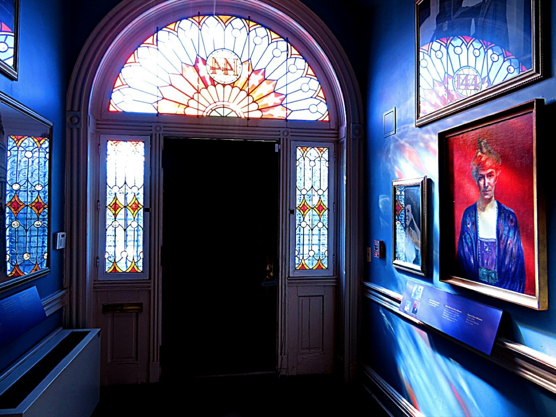 Sunlight streams through stained glass windows into a dark foyer with artwork celebrating women