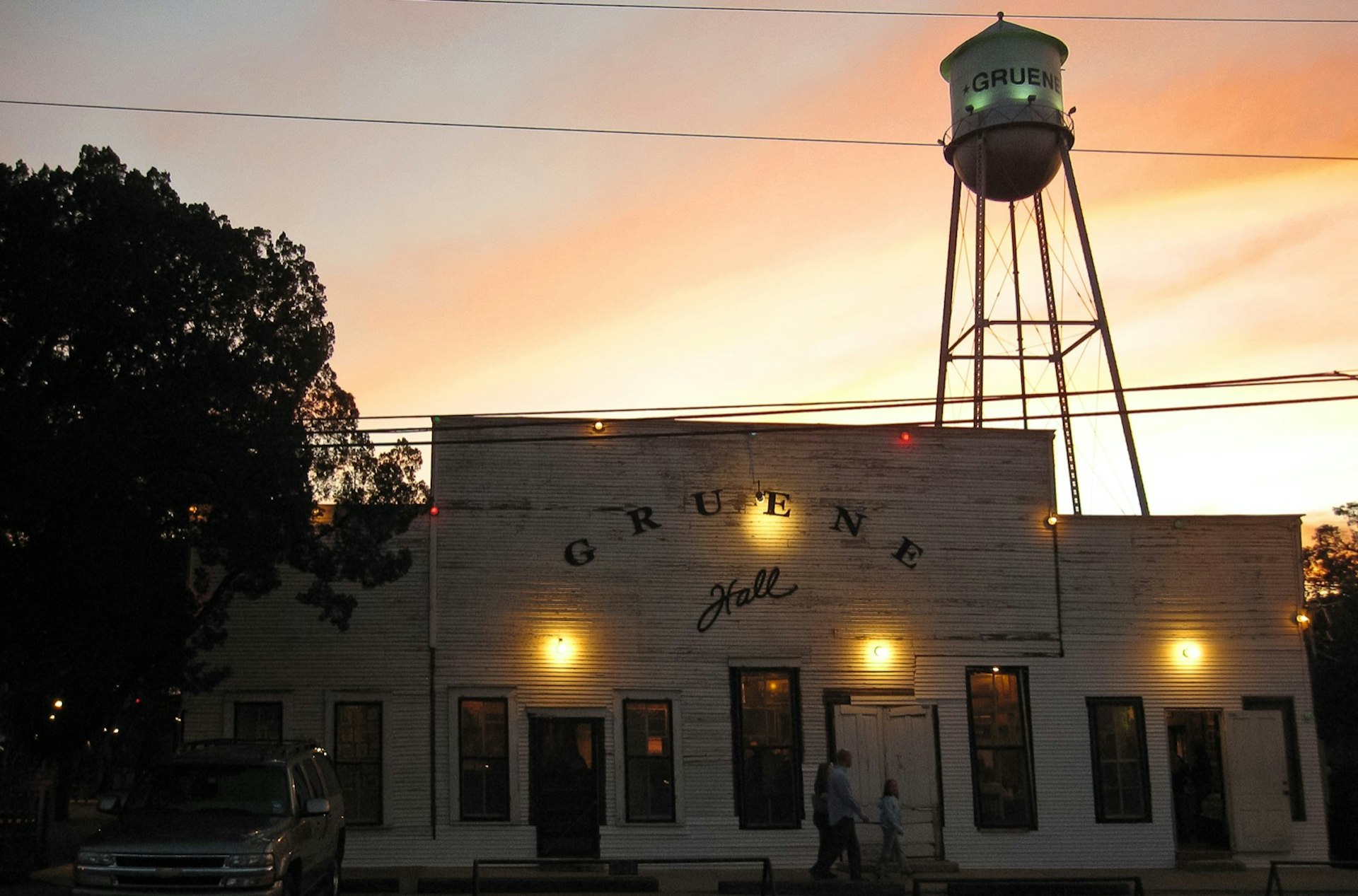 Small amber lights illuminate a whitewashed dance hall at sunset as a water tower looms in the background in the Hill Country