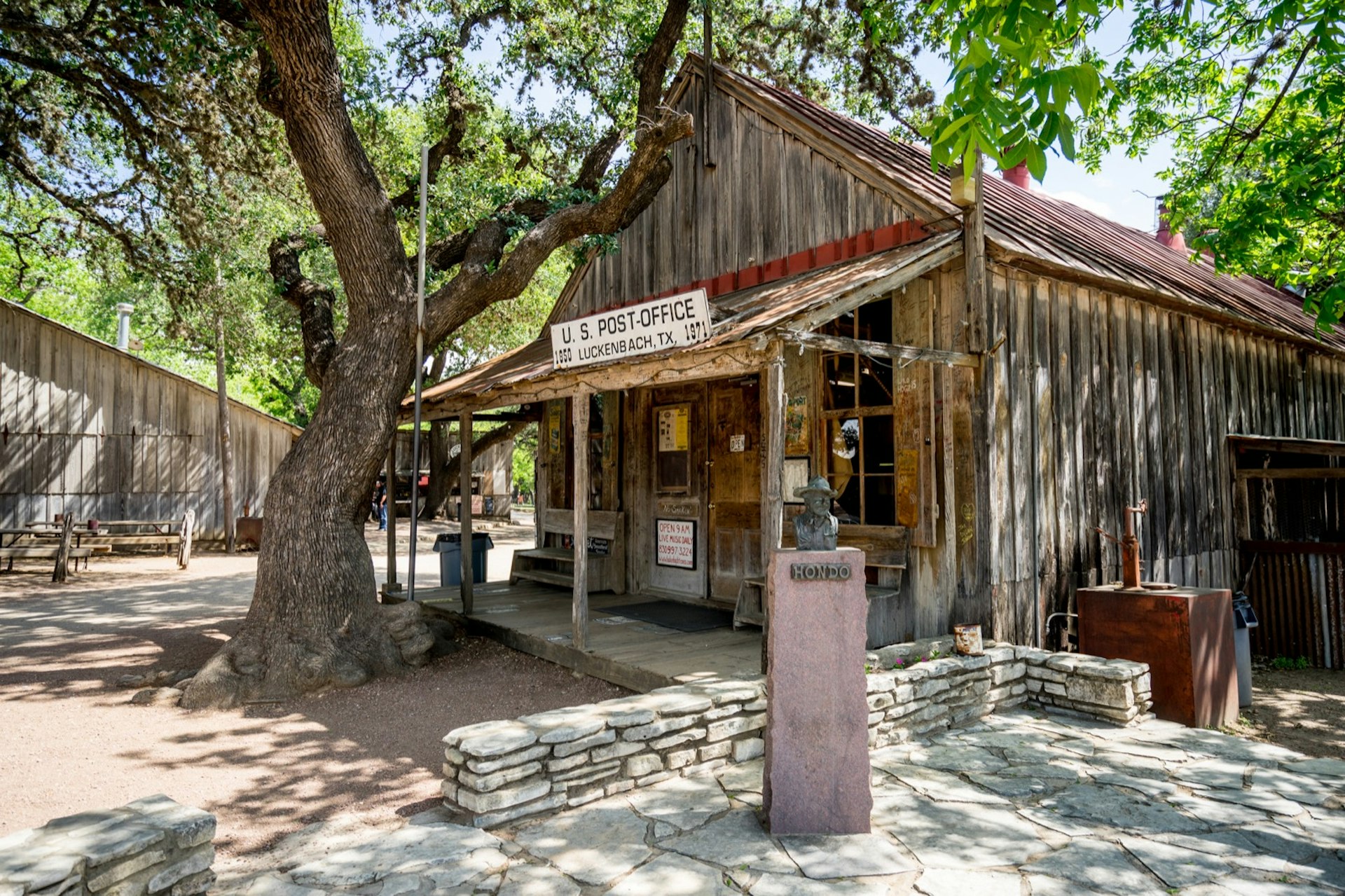 A bust sits outside an old-fashioned looking building with a large tree encroaching on it in the Hill Country