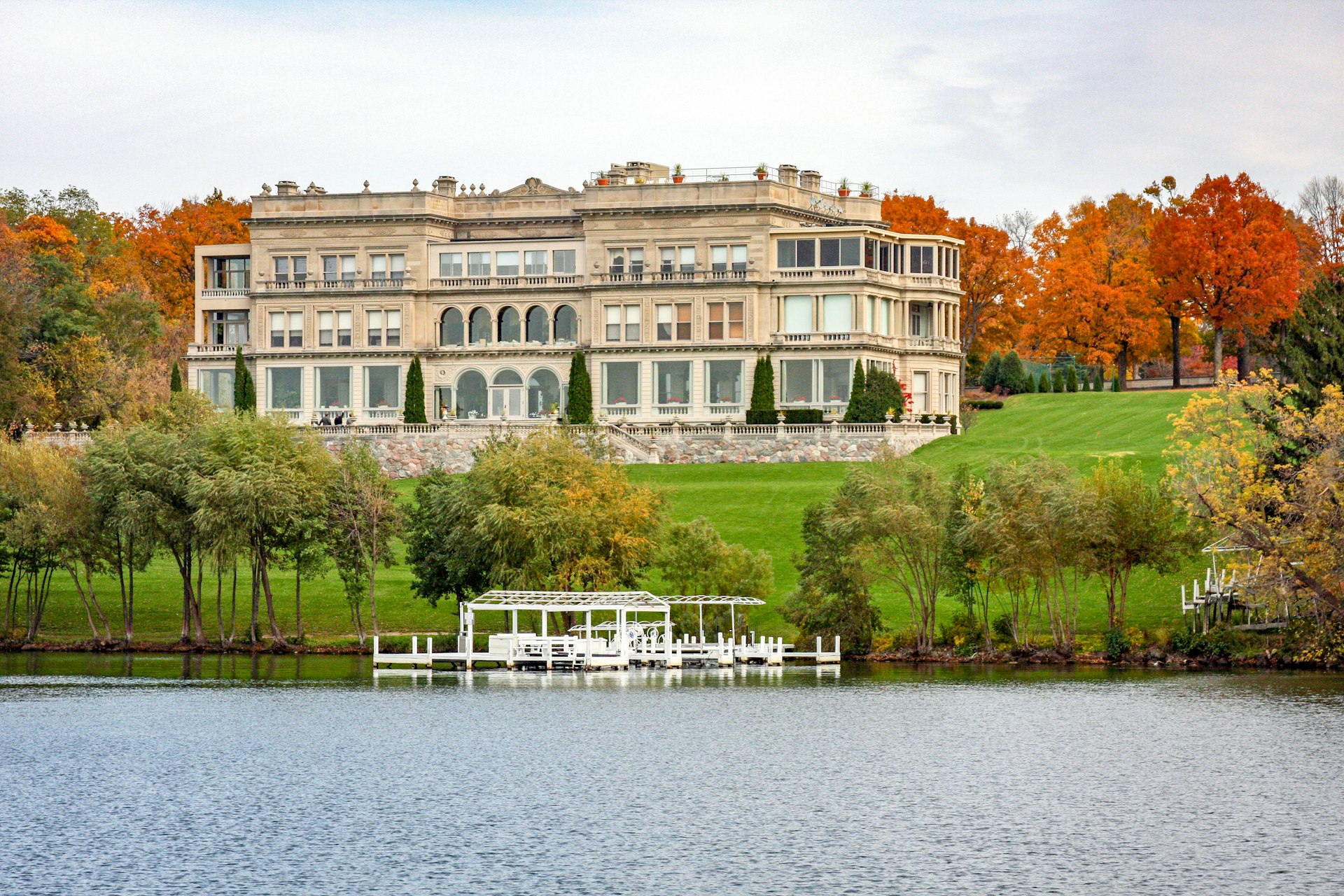 A large mansion sits on a green hill backed by orange and red trees, with a small dock and Lake Geneva in the foreground
