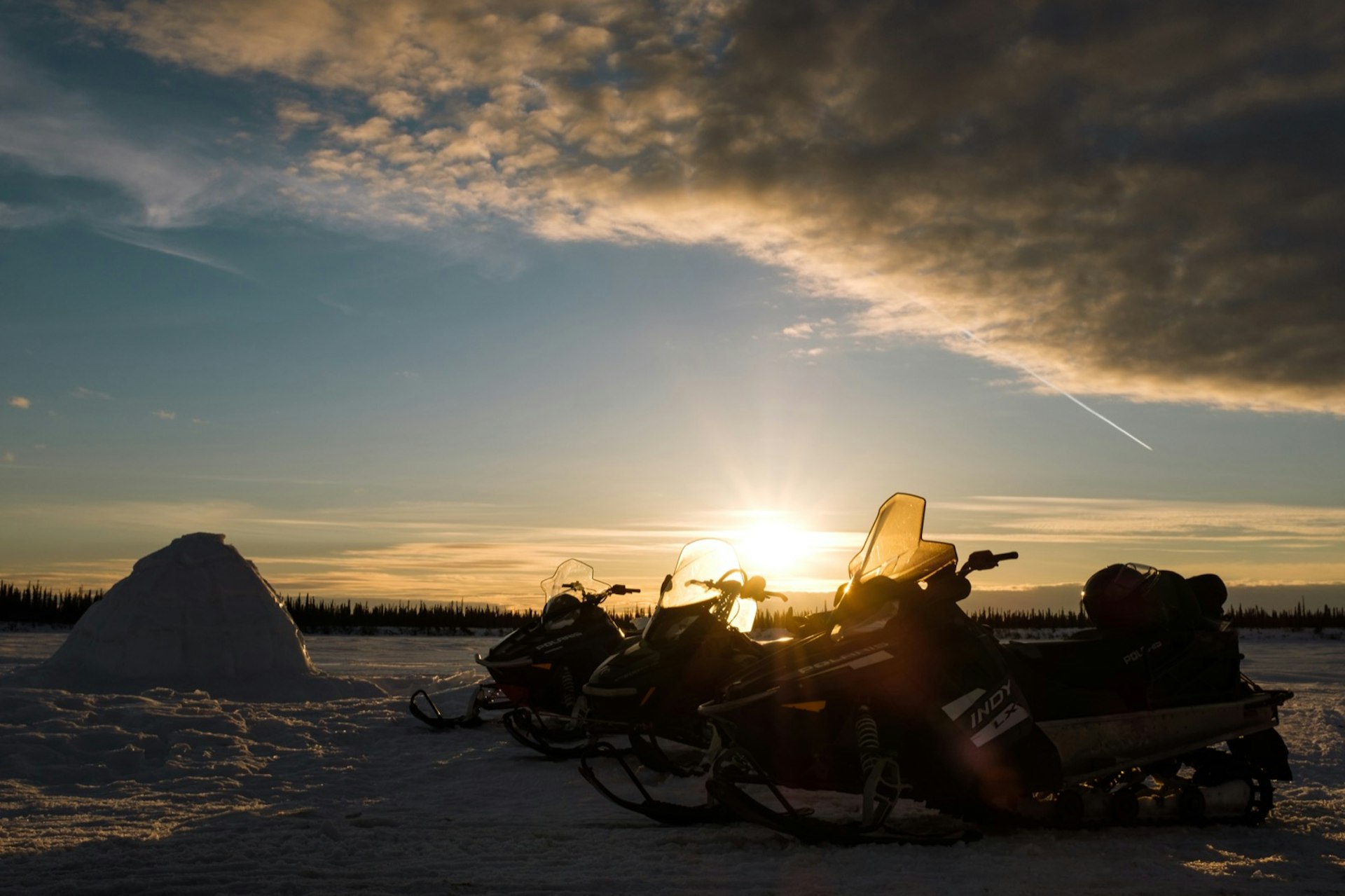 The sun goes down behind a row of snowmobiles parked next to an igloo in the Northwest Territories