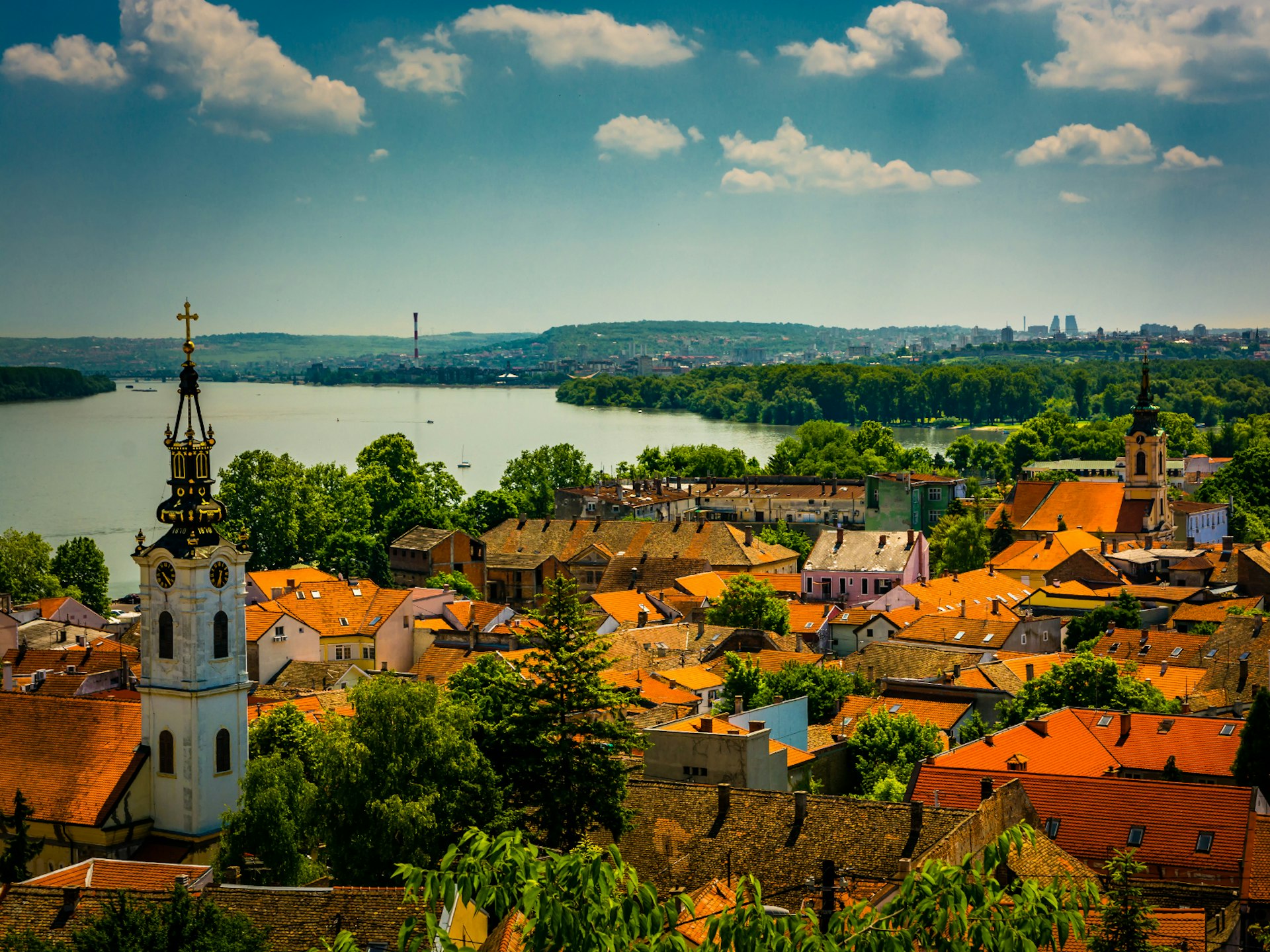 View over the orange rooftops of the Zemun district of Belgrade; there are also trees, a church with a black and gold spire, and the River Danube beyond.