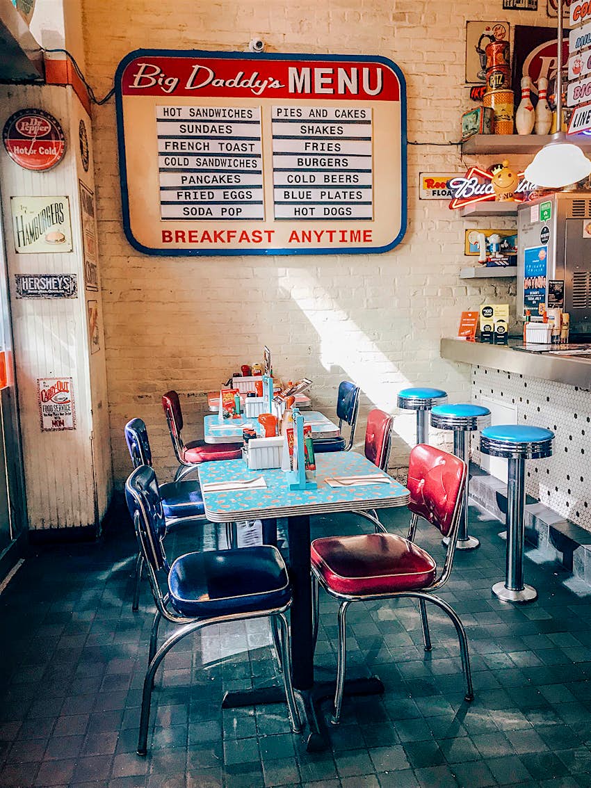 A view of a table with blue and red vinyl chairs sitting in front of a menu board at Big Daddy's in New York City