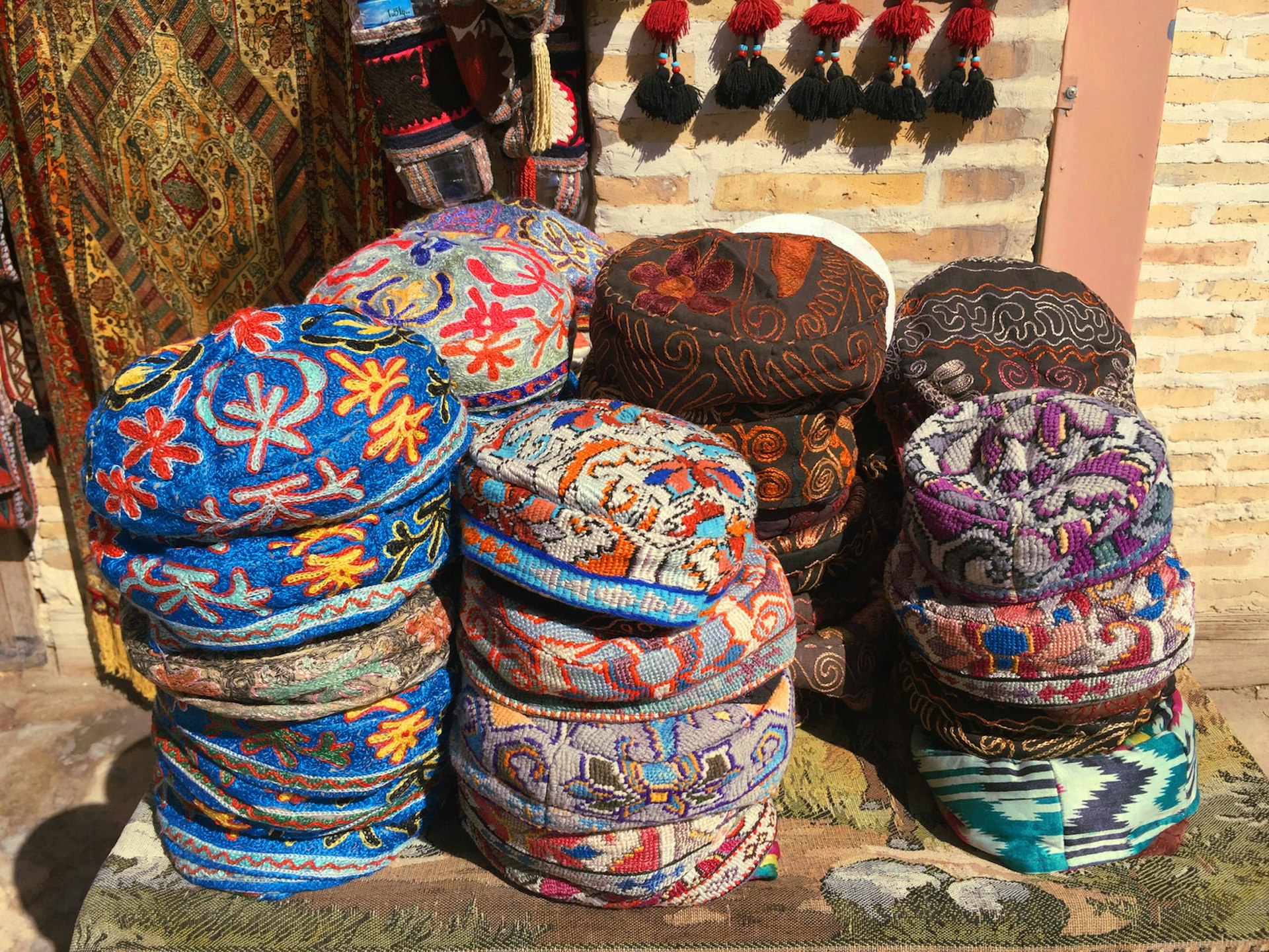 Stacks of brightly coloured embroidered round hats on a small table in front of a brick wall