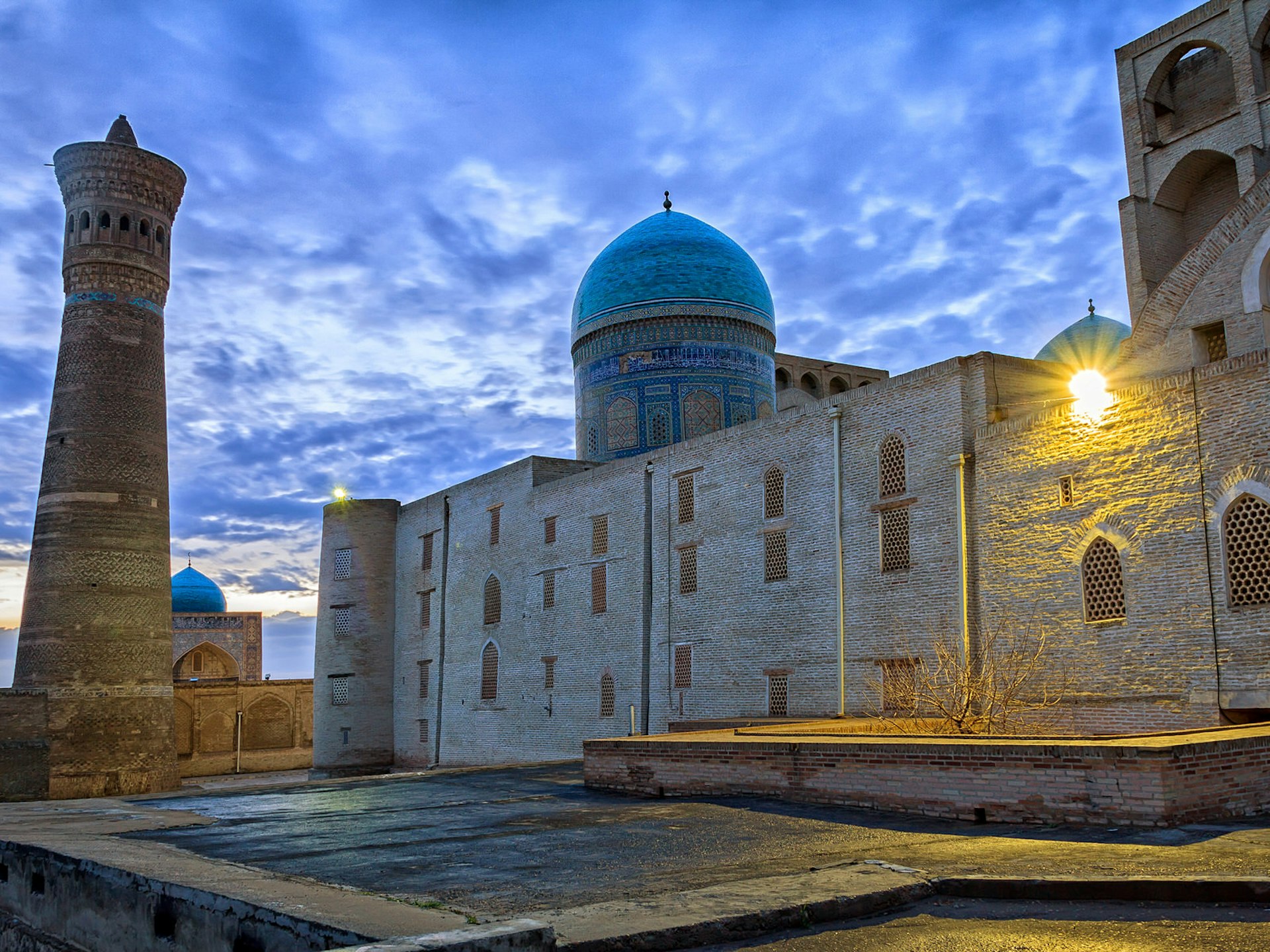 The blue dome and brown mud structures of the Kalon Mosque and minaret under cloudy, dusk skies.