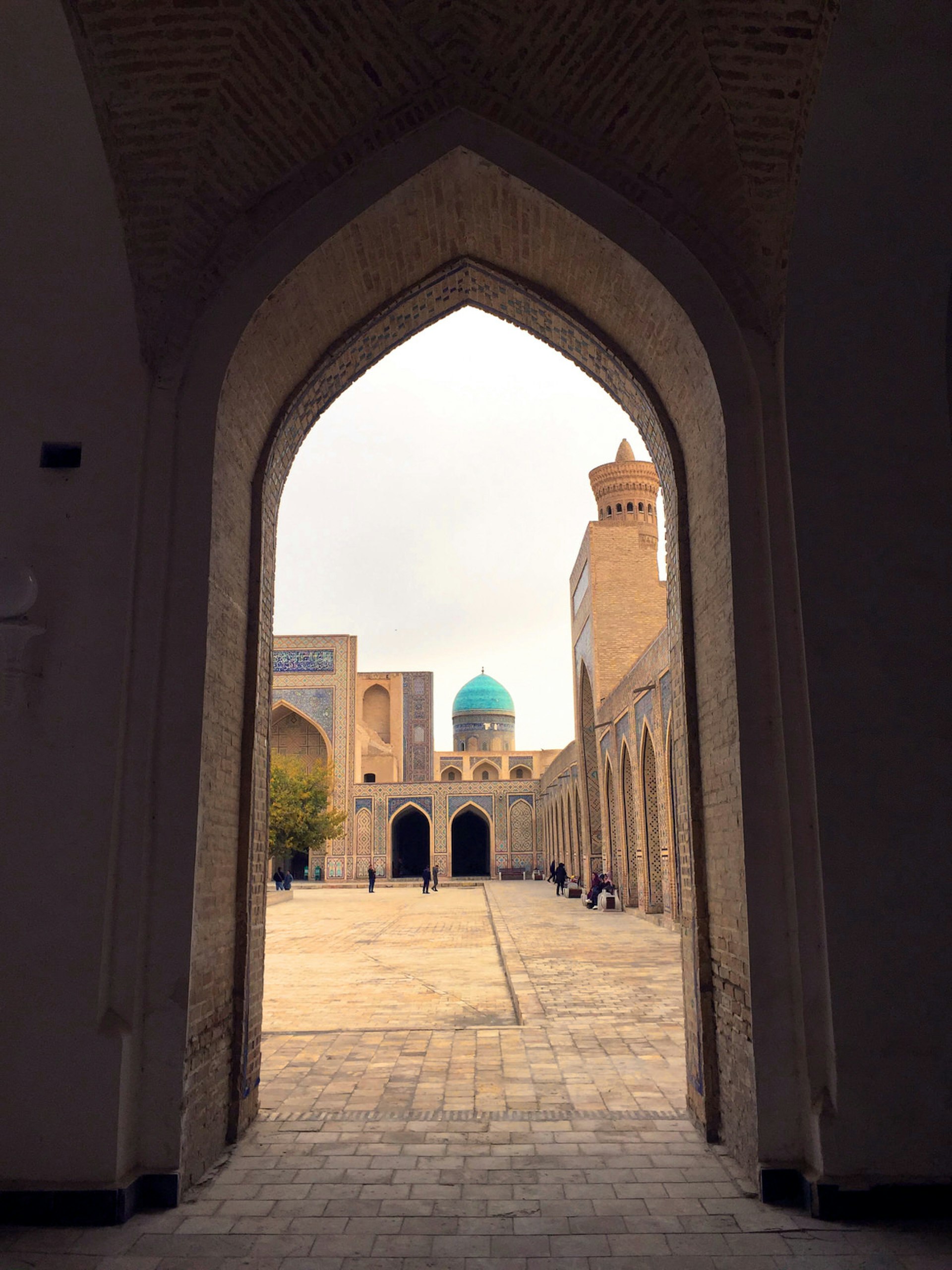 The inner courtyard of Kalon Mosque with blue dome and minaret as seen through a pointed arched doorway.