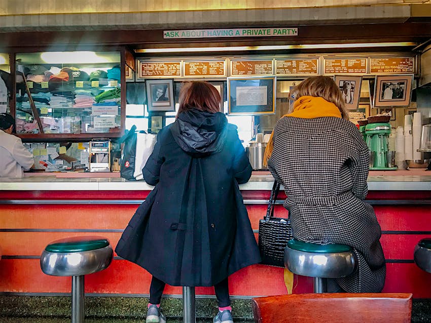 Two women wearing coats sit on vinyl-covered stools at a diner counter in New York City