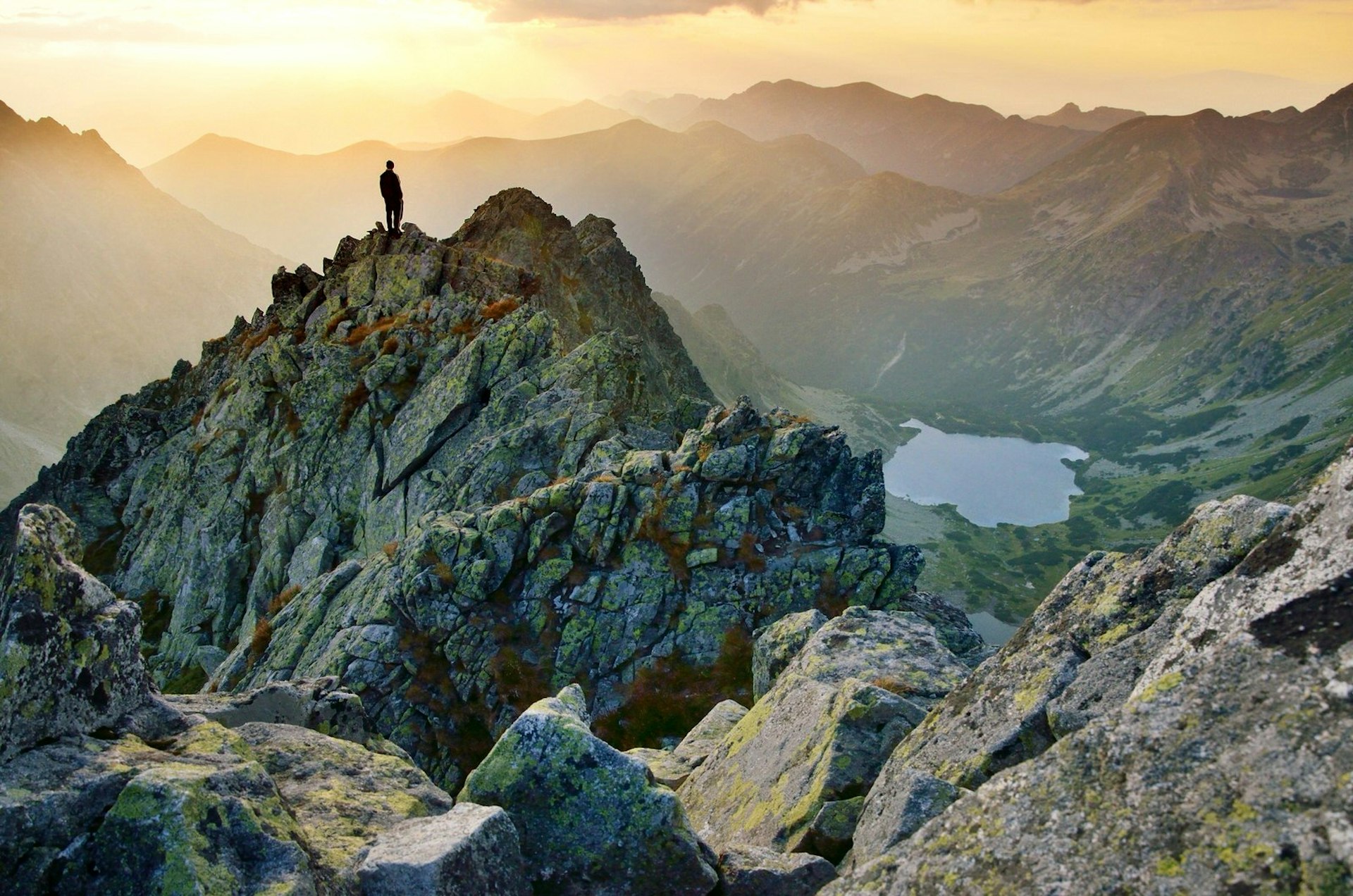 A man stands on top of a jagged peak in the High Tatras, Slovakia, looking down on a mountain lake at sunset