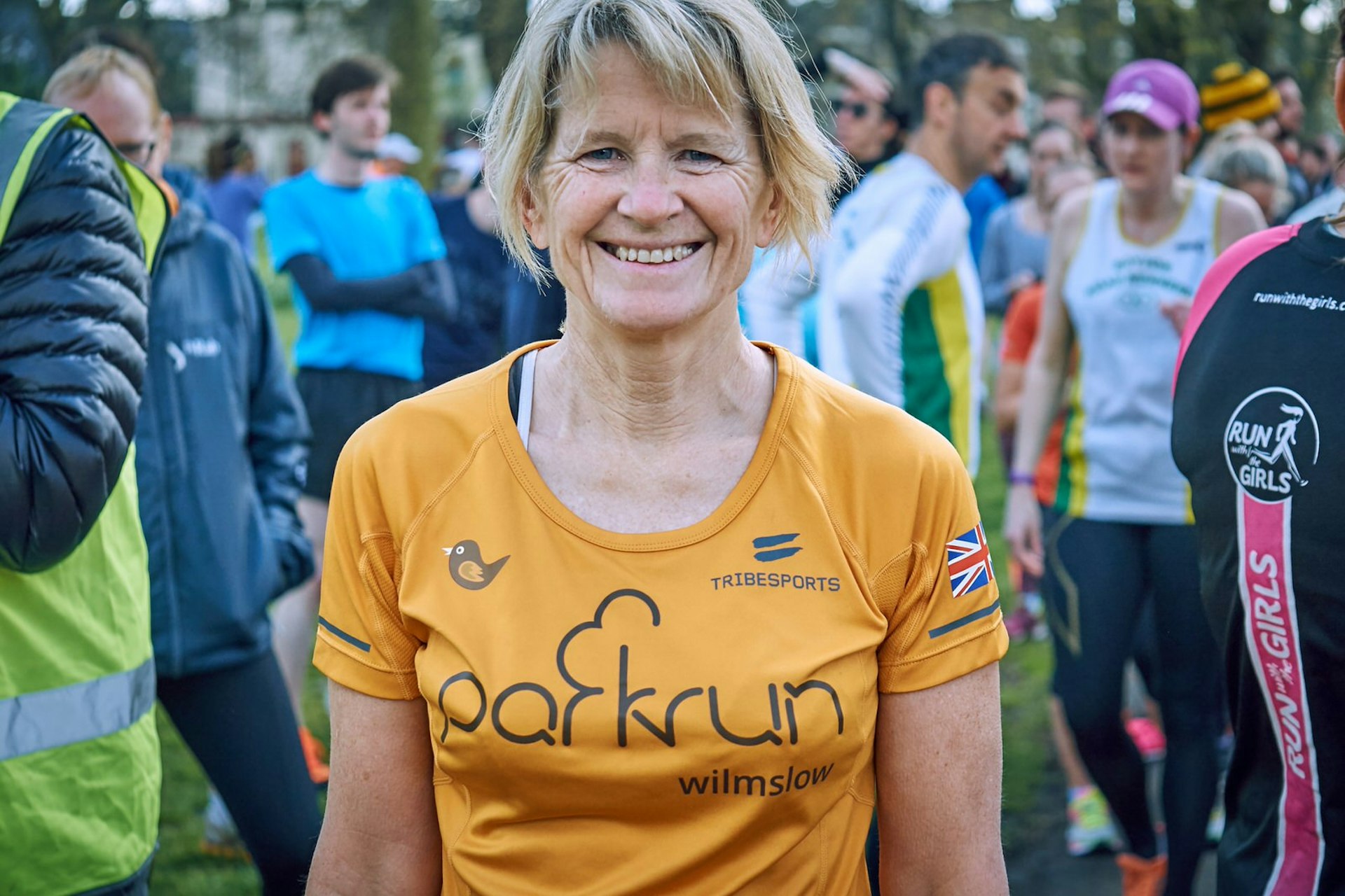 A woman in an orange parkrun shirt looks into the camera with a broad smile on her face; behind her are other runners milling 