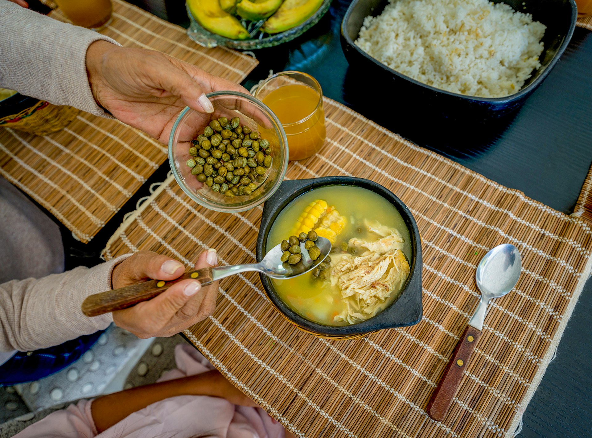 A person adds capers to a bowl of chicken and corn stew. A bowl of rice and a bowl of avocado sit on the table behind it. Colombia.