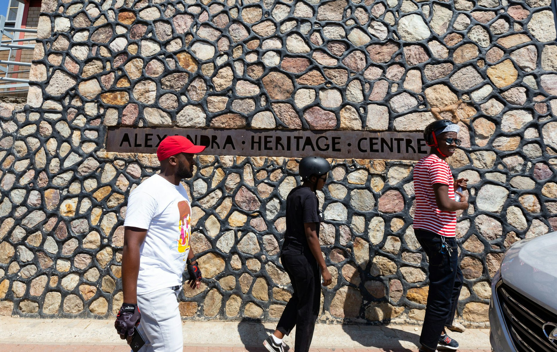 Johannesburg June - Three people, two wearing black bicycle helmets, the other in a red baseball hat, walk in front of a huge stone wall - rounded cobbles of pinkish stone with dark grey mortar - which has the cast iron sign for the Alexandra Heritage Centre