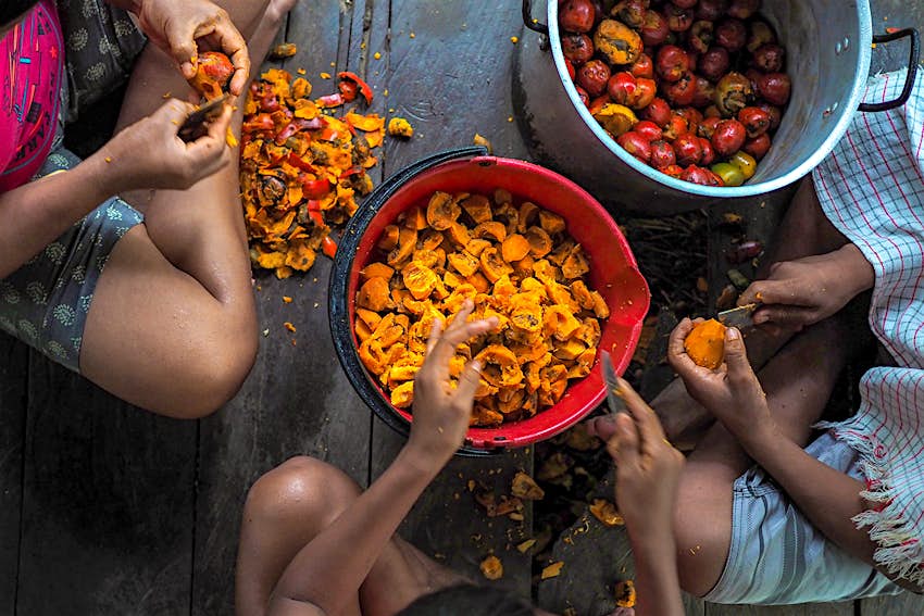 Three people sit around two buckets of orange fruit, peeling off the skins. Leticia, Colombia.