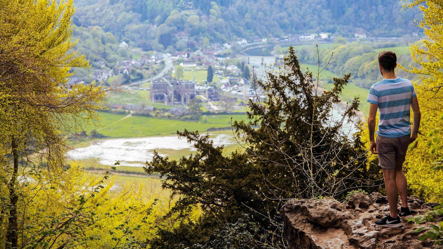 A man stands on a rocky ledge looking down on the ruins of Tintern Abbey