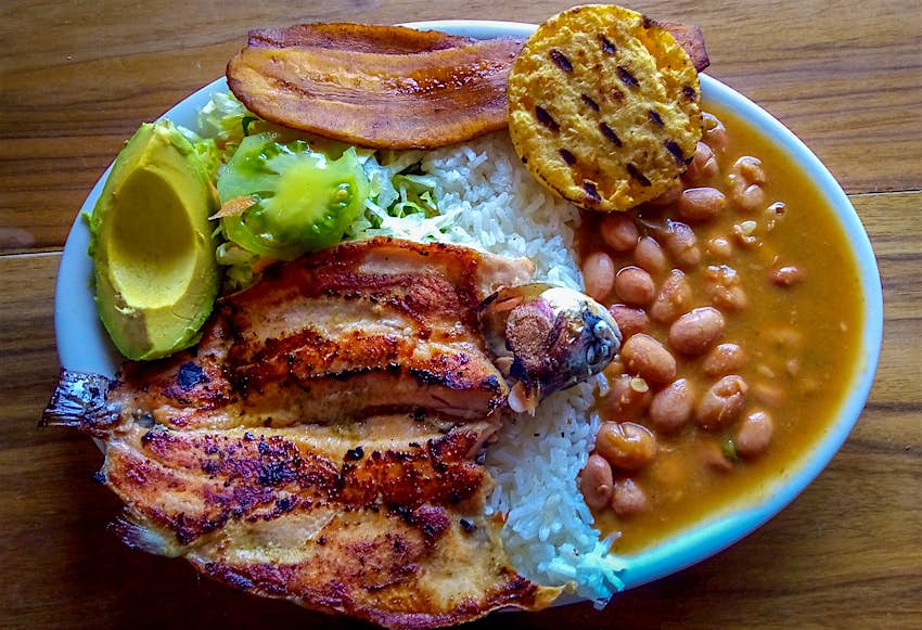A plate with grilled fish, avocado, plaintains, beans and an arepa, all on a bed of white rice. Colombia.