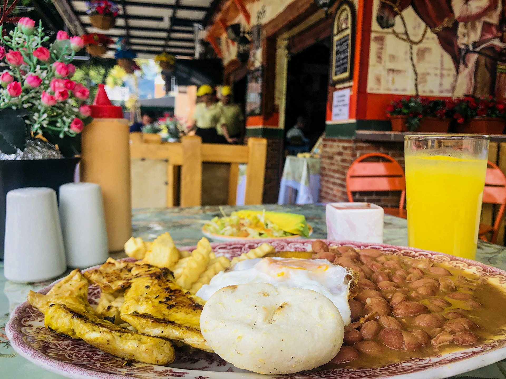 A side view of a plate filled with beans, eggs, fries and arepas and a glass of yellow juice. The background is a colorful restaurant scene in Medellín, Colombia.