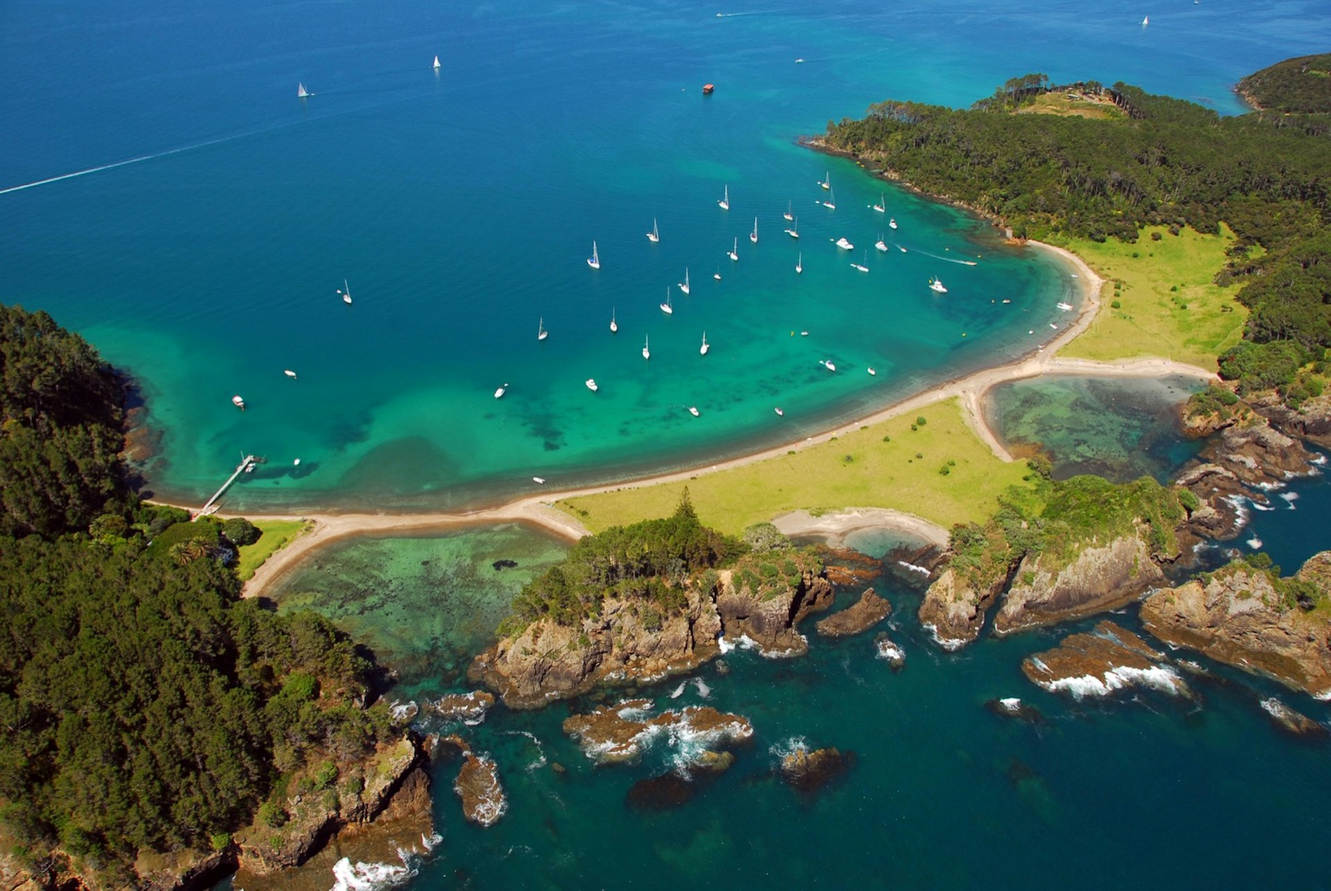 Aerial shot of boats off Roberton Island, Bay of Islands, New Zealand; the boats are all at achor in a horseshoe shaped bay with torquoise waters near the beach it its heart; the ends of the bay are both forested; the opposite side to the bay across a narrow isthmus is a shore dominated by tall rocky outcrops topped by trees