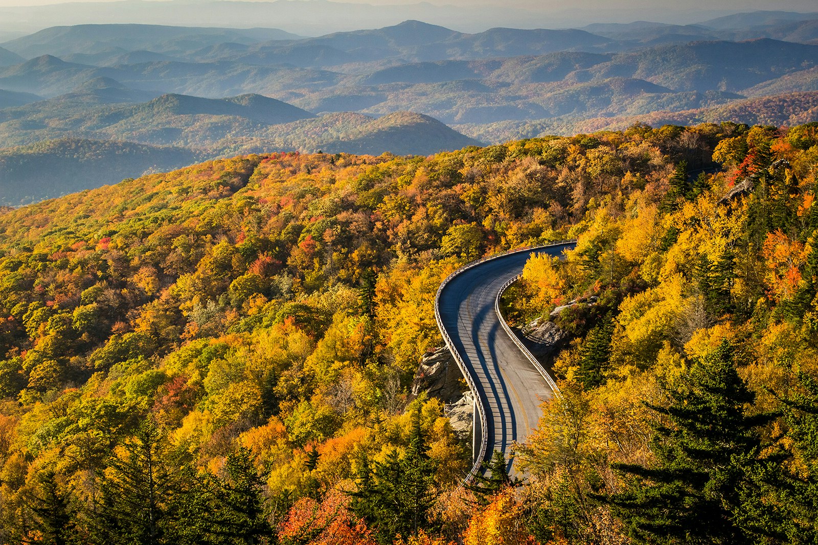 A road winds through trees, with the Blue Ridge Mountains stretching into the distance. The leaves are starting to turn golden. Appalachia, USA.