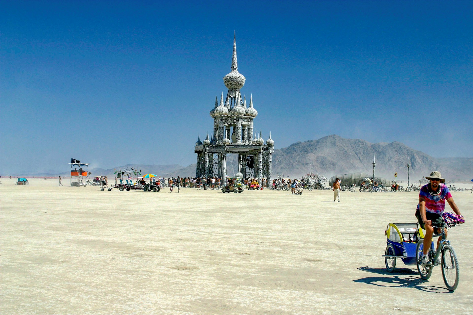 A flat expanse of the Black Rock Desert in Nevada stretches toward a mountain range as people gather at an art installation styled like a building with minarets. A man on a bike pedals in the foreground.