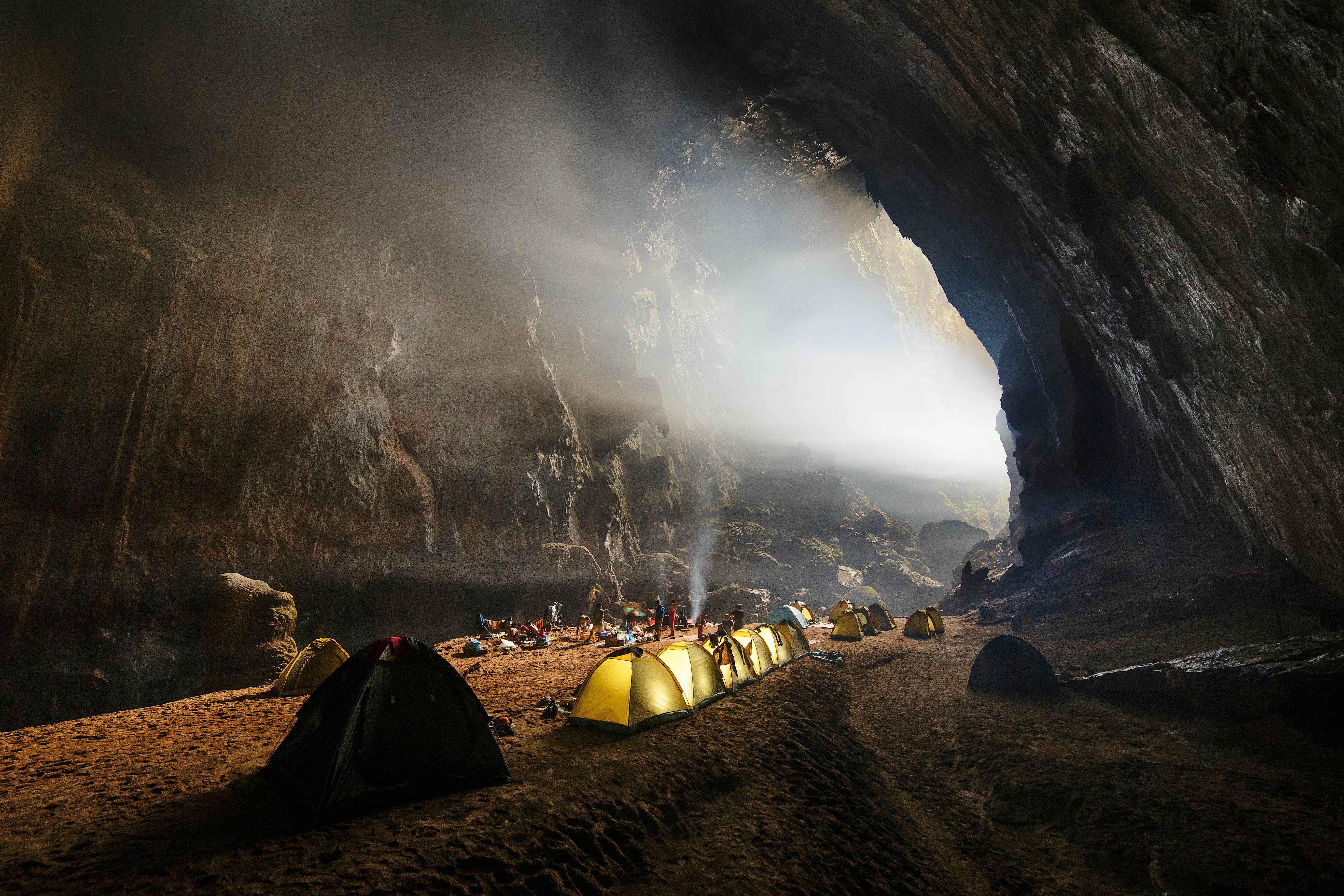 A row of tents is lined up on the rock in the cave, while sunlight beams in through an opening.