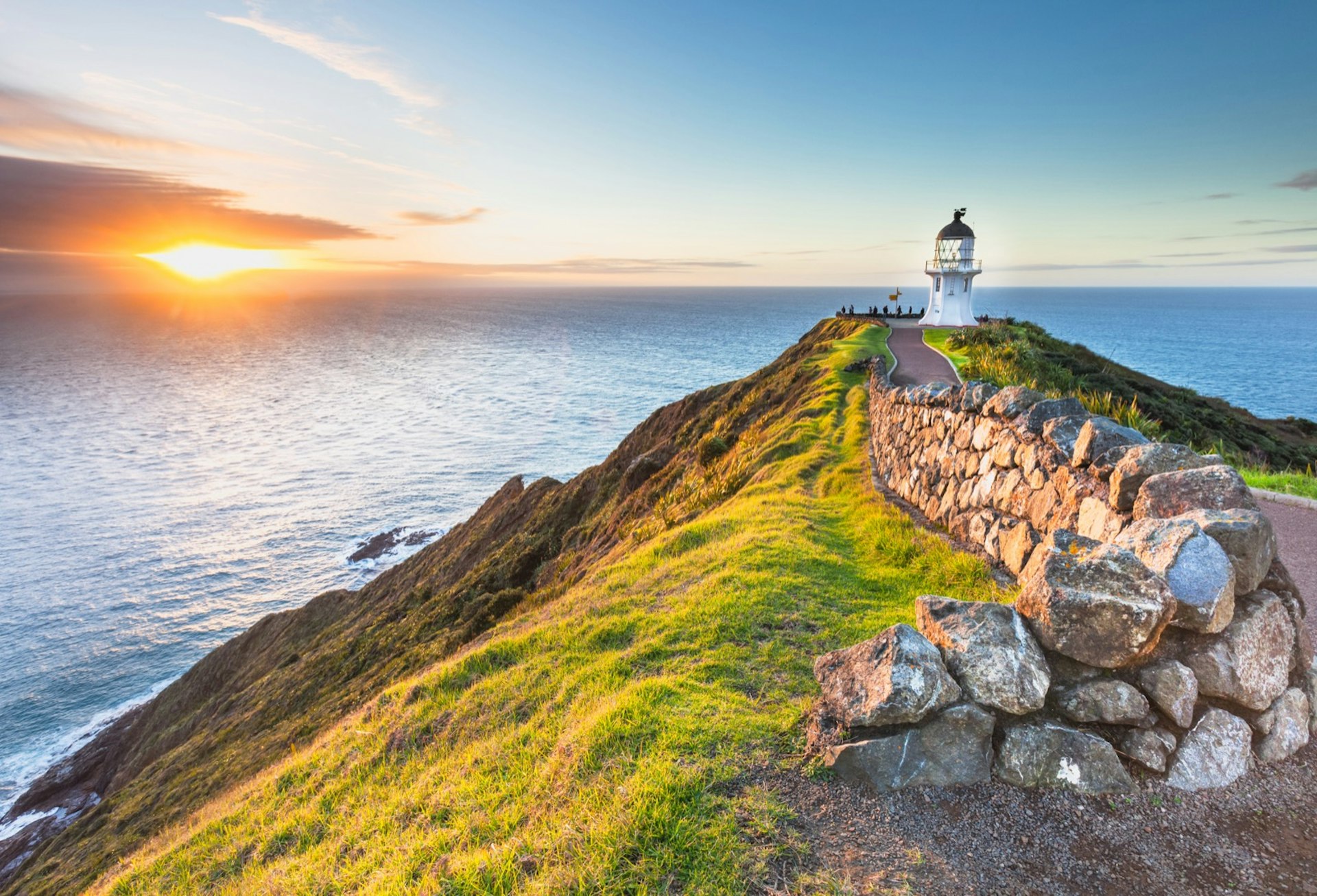 A golden sun pierces a dark cloud on the horizon as it sets into the ocean, the rest of the sky is various hues of beautiful blue; in the foreground is a rocky Cape Reinga, with its narrow flat top lined with grass and a stone wall that flanks a paved path to the Cape Reinga Lighthouse