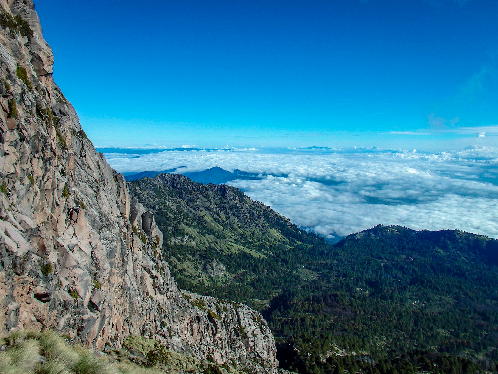 A high-altitude photo of a low, forest-covered valley and a craggy rock face on the summit of Nevado de Colima