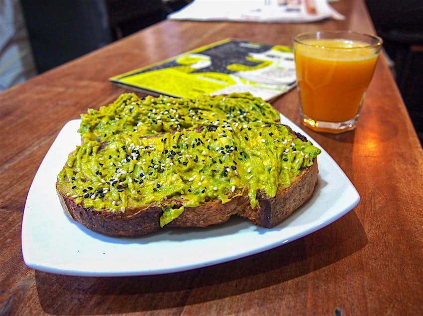 A toast spread with avocado and topped with small seeds, with a small cup of orange juice in the background. Santiago, Chile.