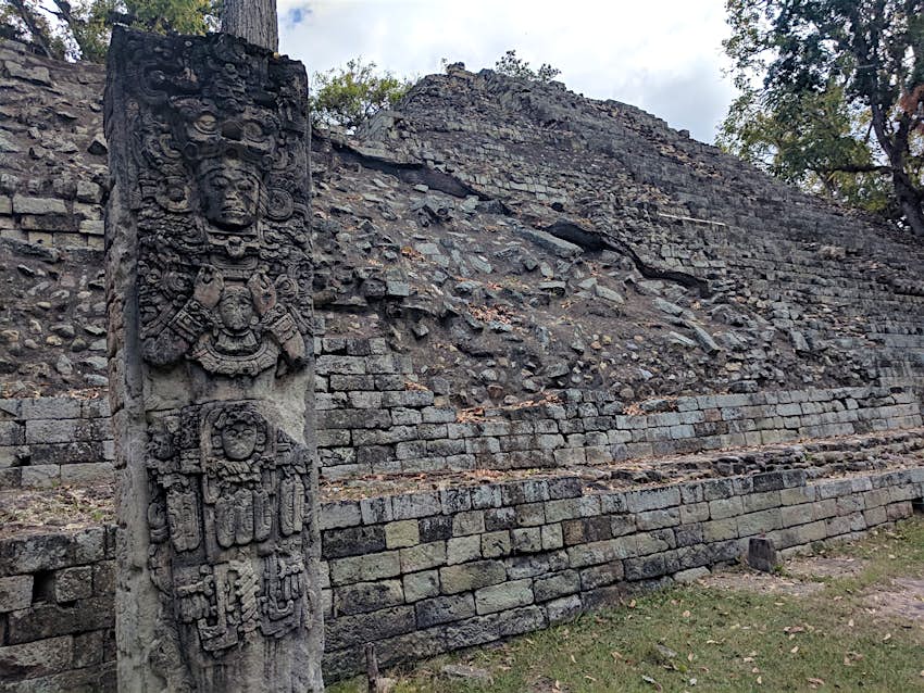 A mayan stone statue full of ornate carvings, thought aged and stained black by weathering, stands in front of crumbling stone pyramid at Copan Ruins in Honduras 