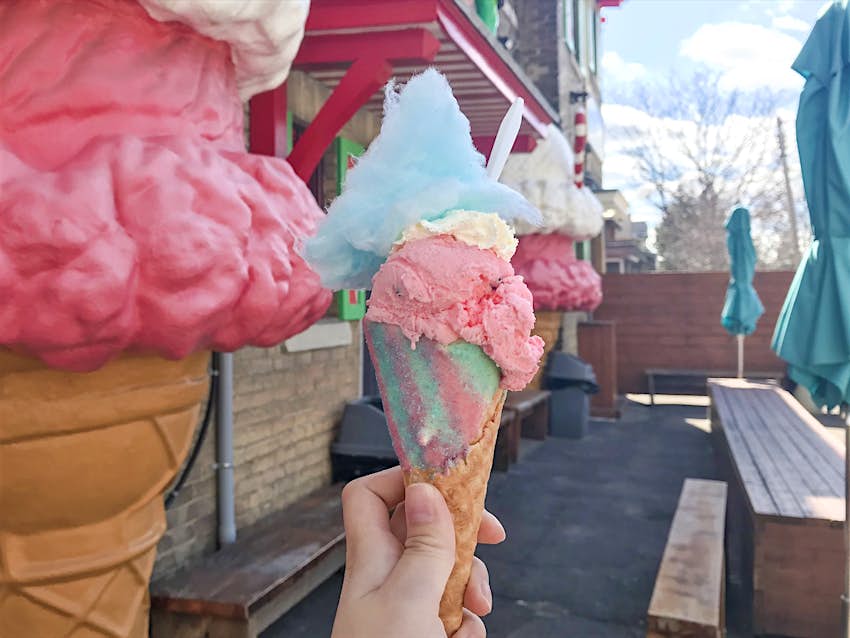 A pink and blue fancy ice cream cone is held in front of giant fiberclass cones at a shop