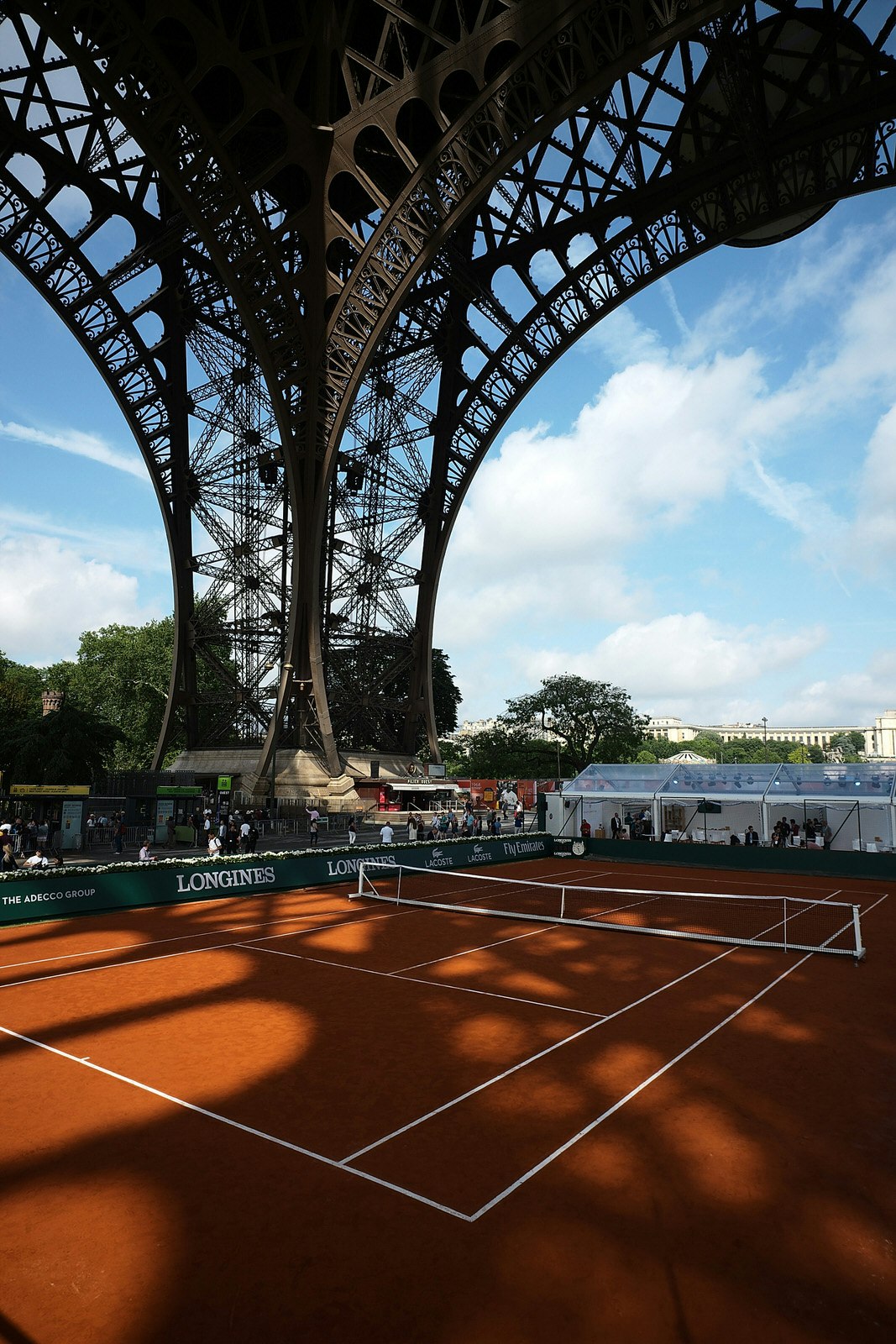 A red clay tennis court, with crisp white lines, sits beneath the elegant iron arches of the Eiffel Tower's large feet; ornate shadows cast by the steel structure spread across the court, with a blue sky in the background