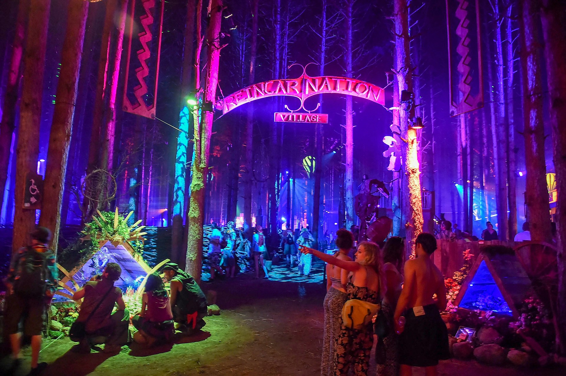 People stand beneath a sign that says Reincarnation village in a forest lit up blue, purple and pink