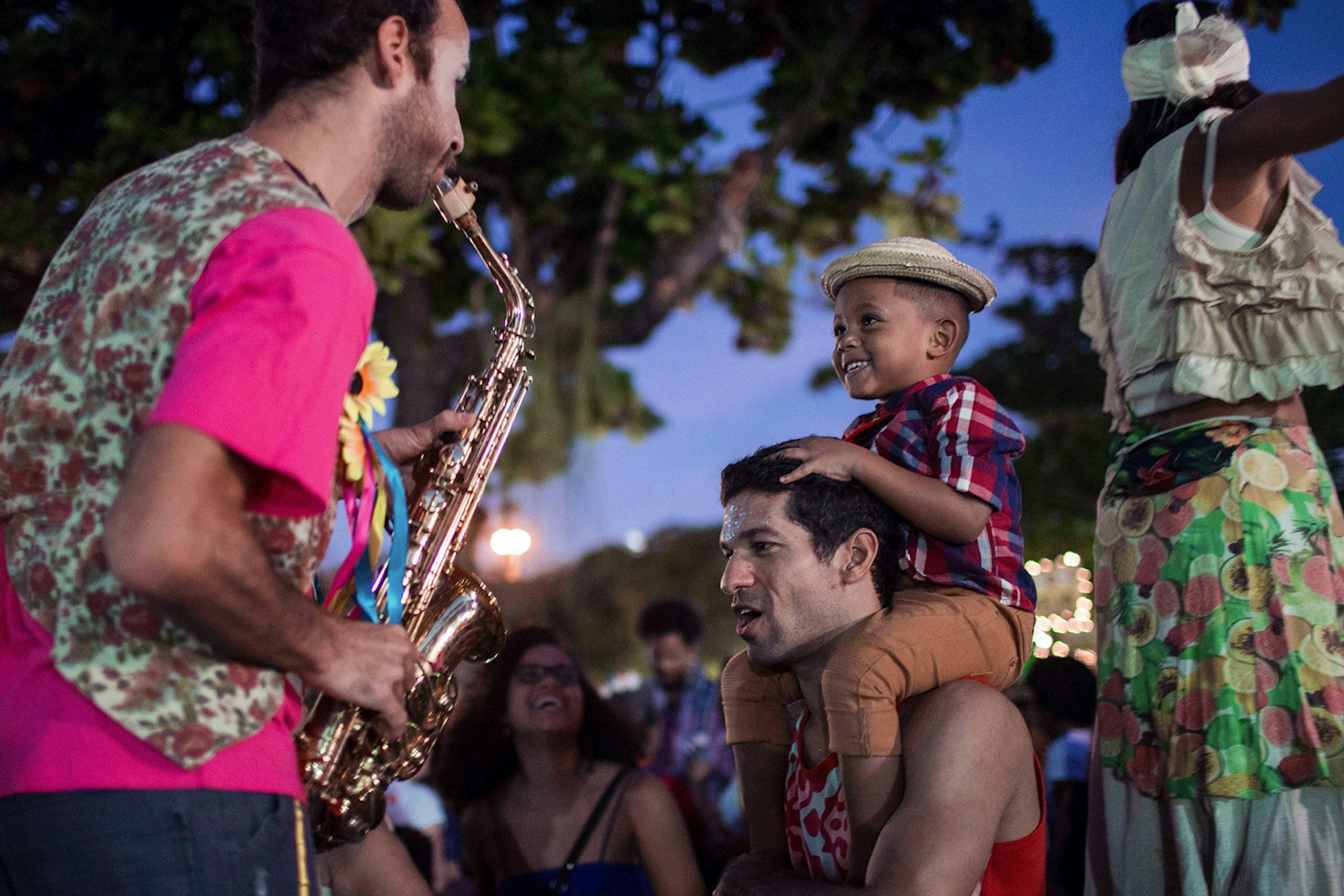 A little boy wearing a hat sits on a man's shoulders while a man in a pink shirt plays the saxophone for him in Rio de Janeiro, Brazil