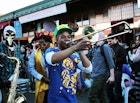 A young man with a bright green baseball hat and blue t-shirt plays a trombone in the street with other revellers, some donning Venetian masks, others feathers in their hair