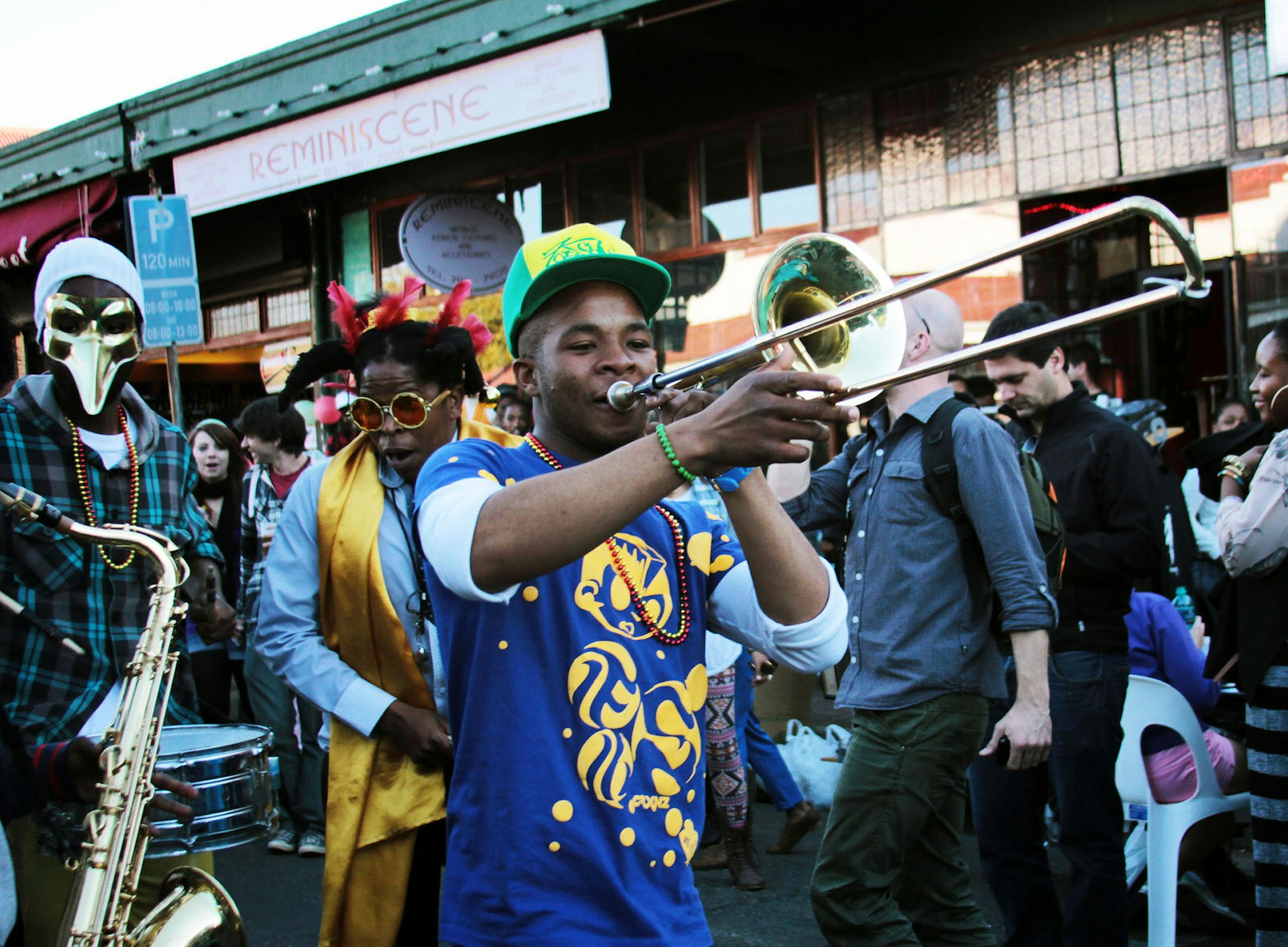 Johannesburg June - A young man with a bright green baseball hat and blue t-shirt plays a trombone in the street with other revellers, some donning Venetian masks, others feathers in their hair