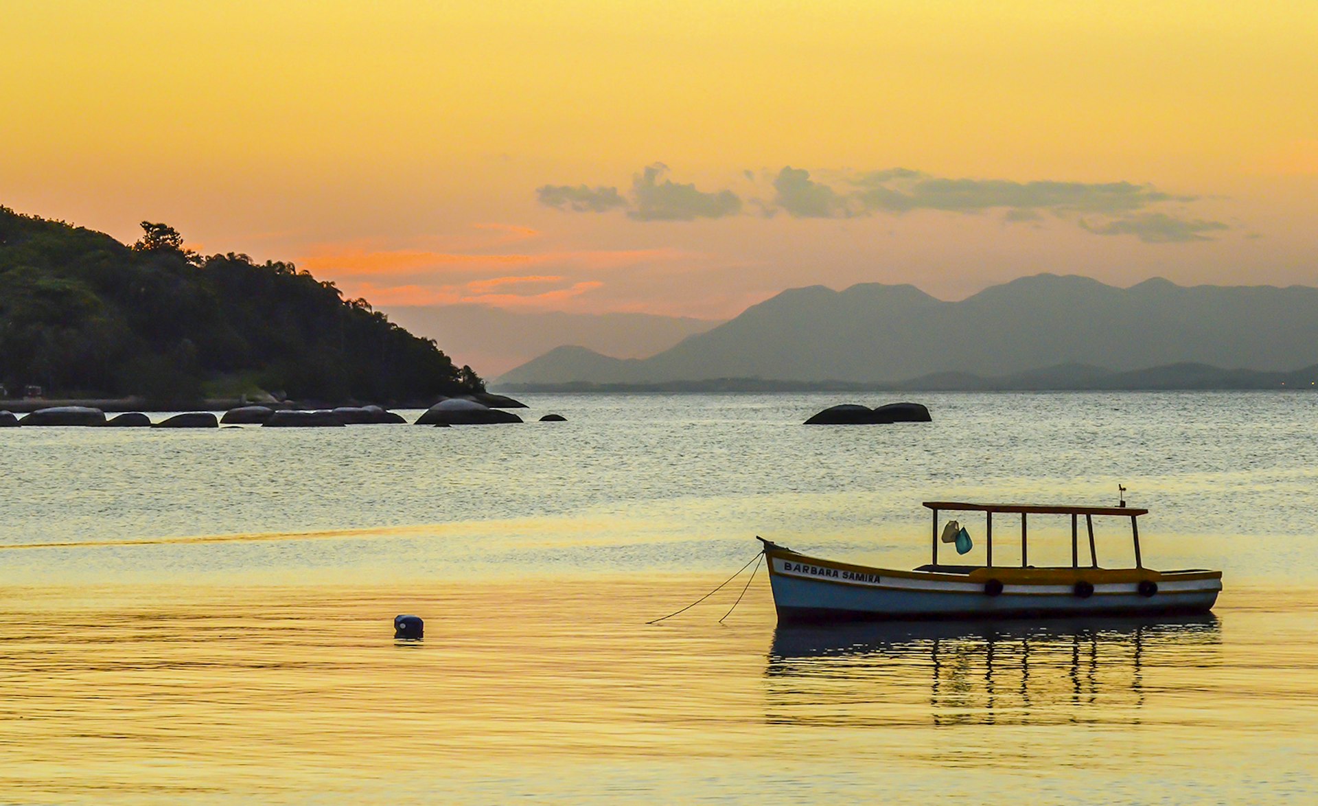 A small fishing boat is moored in glassy water that reflects the orange sky during sunset. Ilha da Paquetá, Brazil.