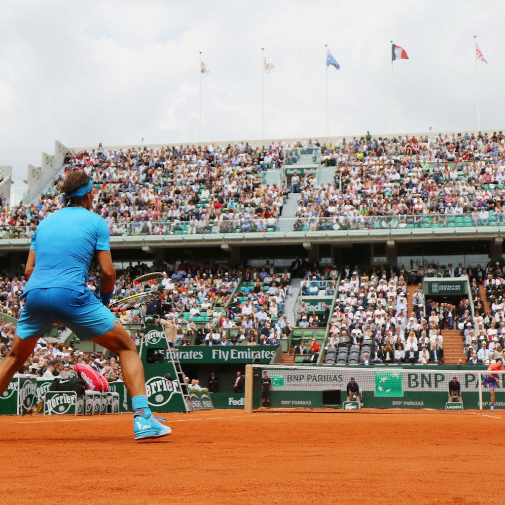 Rafael Nadal, clad in bright blue and crouched in anticipation, awaits a ball served from his opponent across the net; the image is shot from ground level looking up towards Nadal from the back and across to the stands beyond