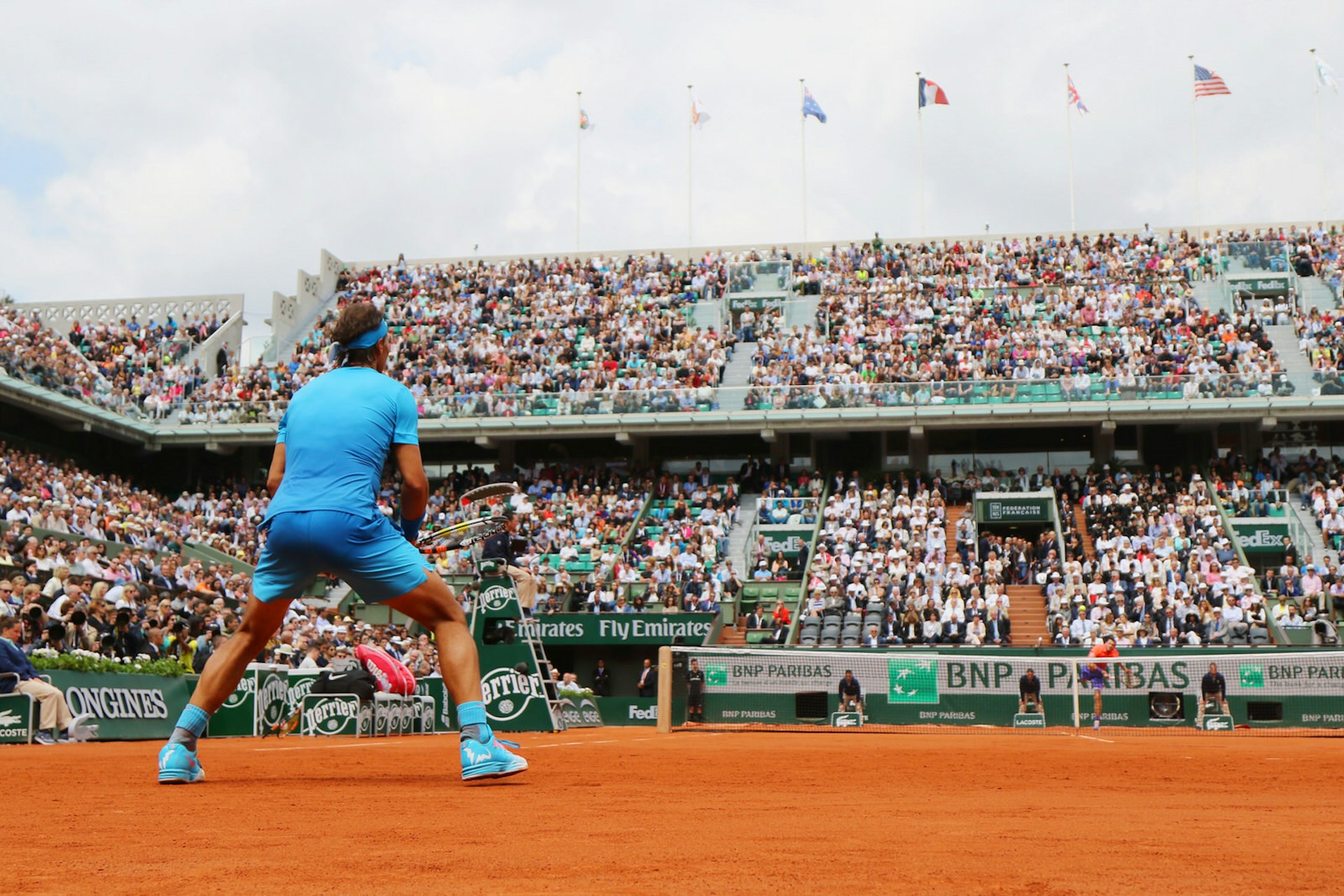 Rafael Nadal, clad in bright blue and crouched in anticipation, awaits a ball served from his opponent across the net; the image is shot from ground level looking up towards Nadal from the back and across to the stands beyond