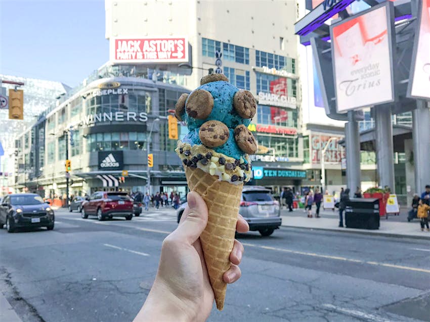Full mini chocolate chip cookies are perched on two scoops of blue ice cream in a cone held at a busy intersection