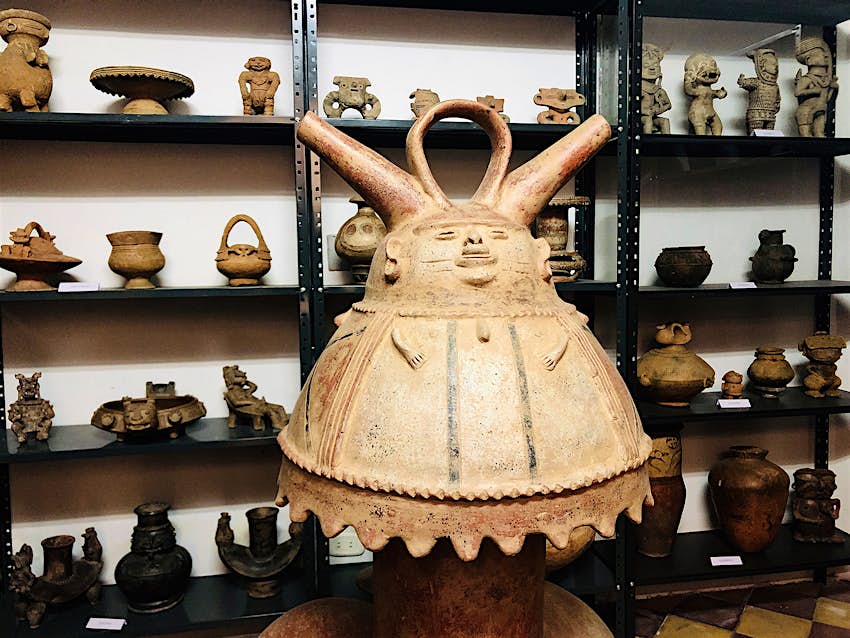 A large light-colored, bell-shaped artifact in the likeness of a big-bellied man is positioned in front of three selves containing a collection of smaller artifacts in Medellín, Colombia. 