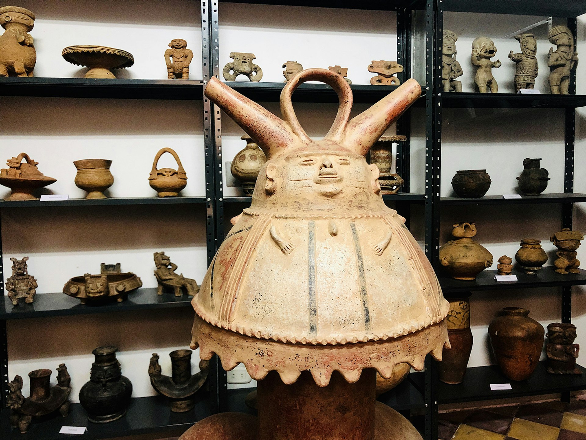 A large light-colored, bell-shaped artifact in the likeness of a big-bellied man is positioned in front of three selves containing a collection of smaller artifacts in Medellín, Colombia. 