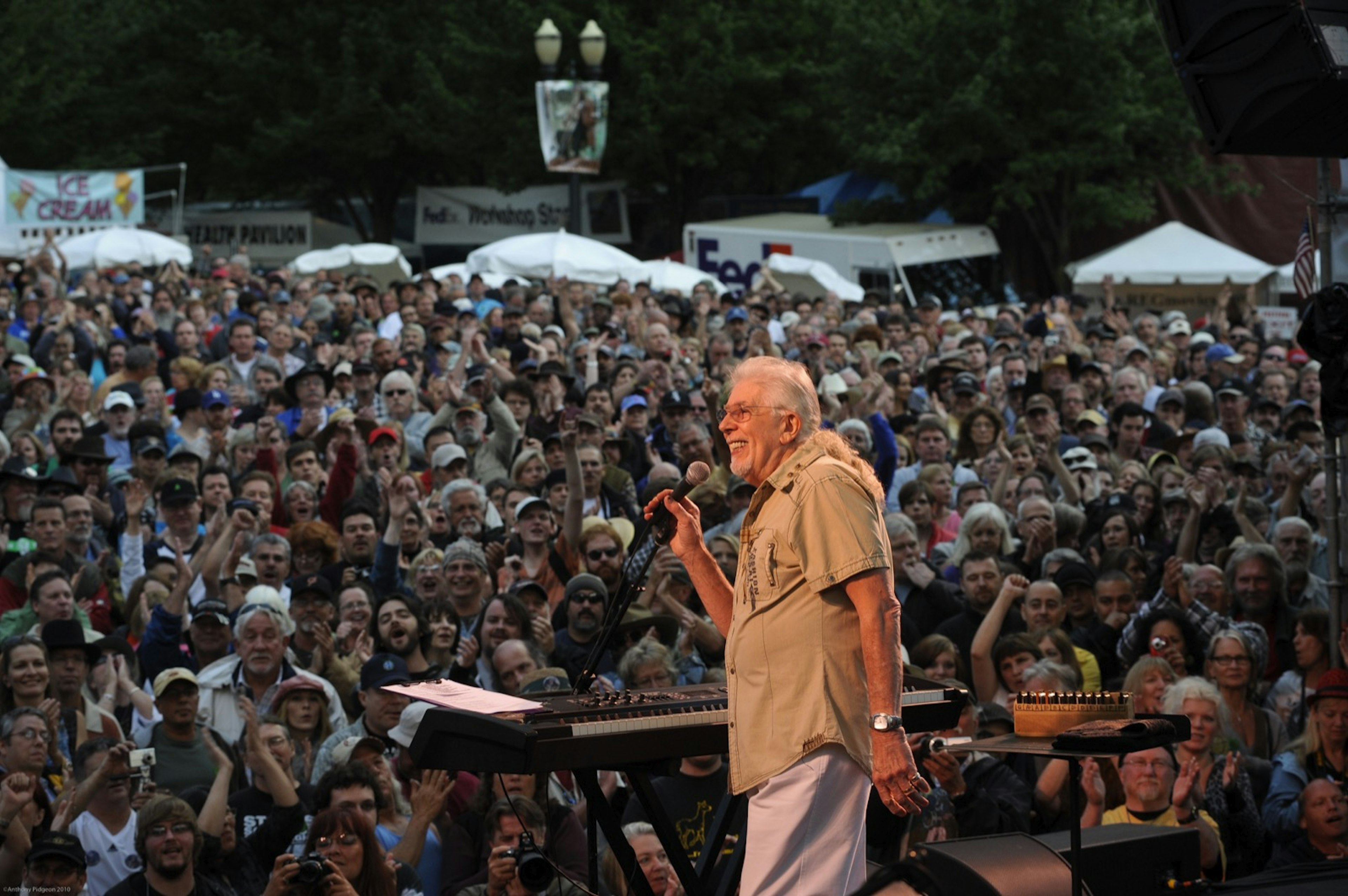 A huge crowd of concertgoers are seen behind a white haired man playing on stage