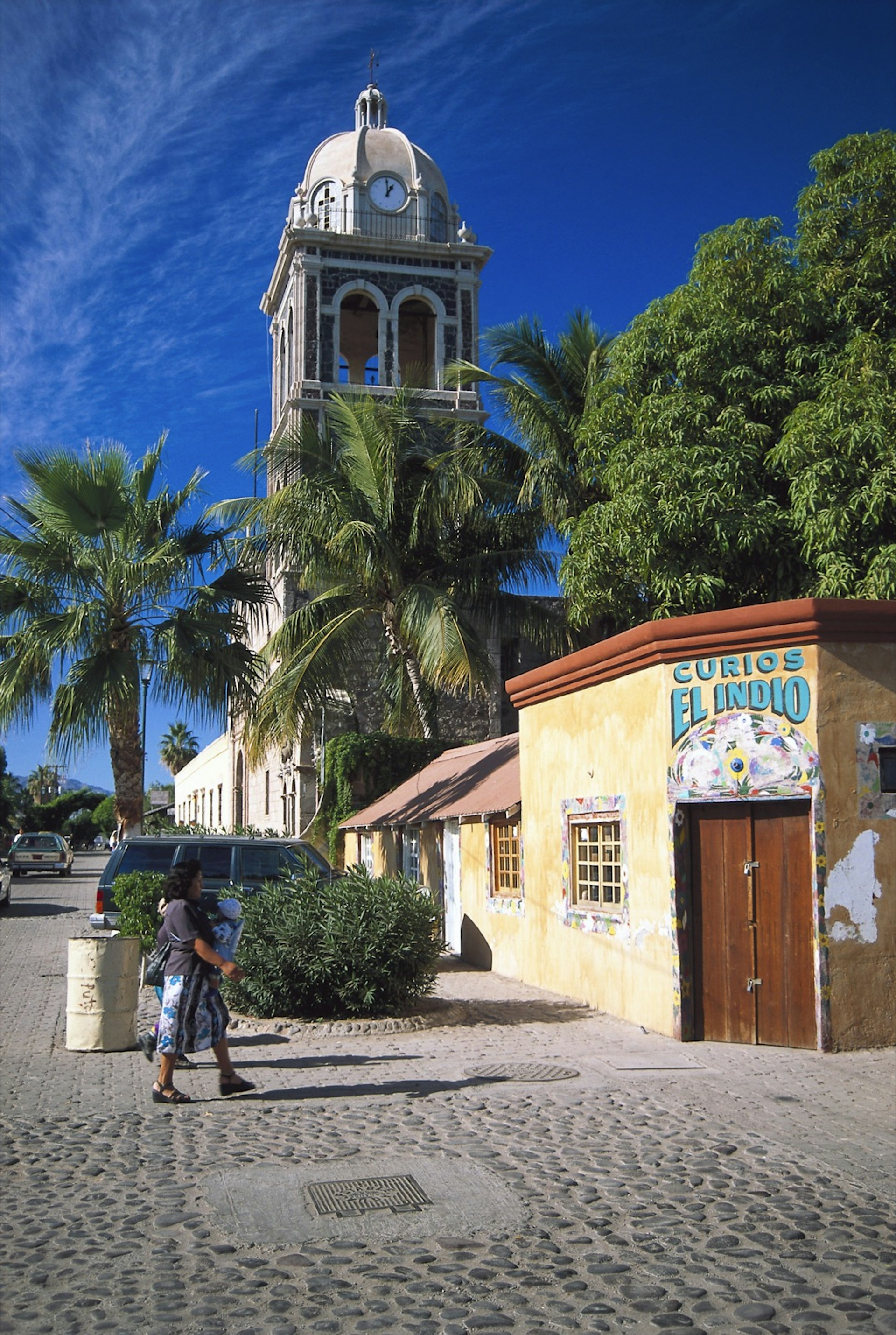 A woman crosses a cobble stone street with a church and palm trees in the background