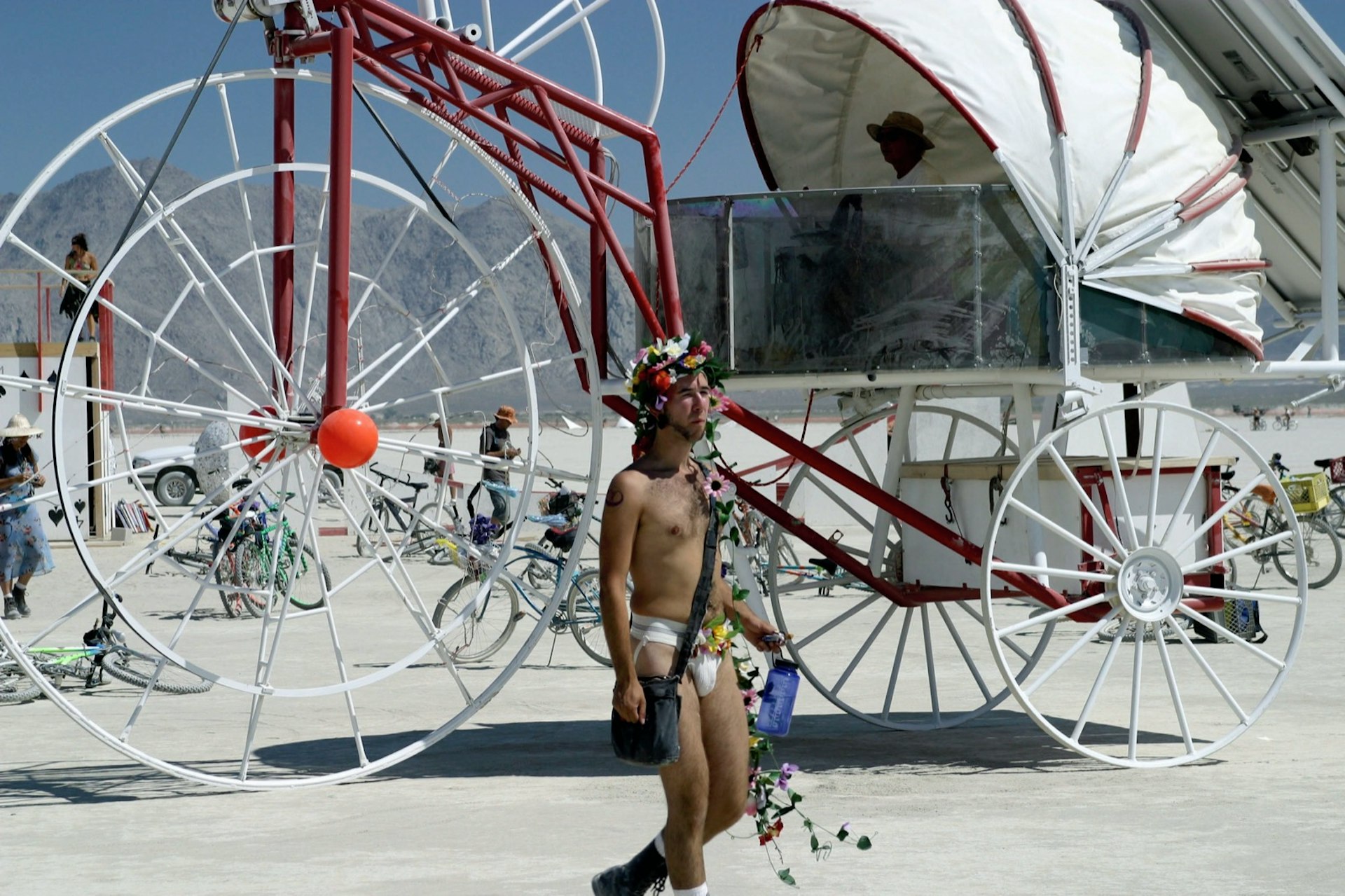 a man wanders past a giant carriage wearing underwear and draped in flowers carrying a water bottle