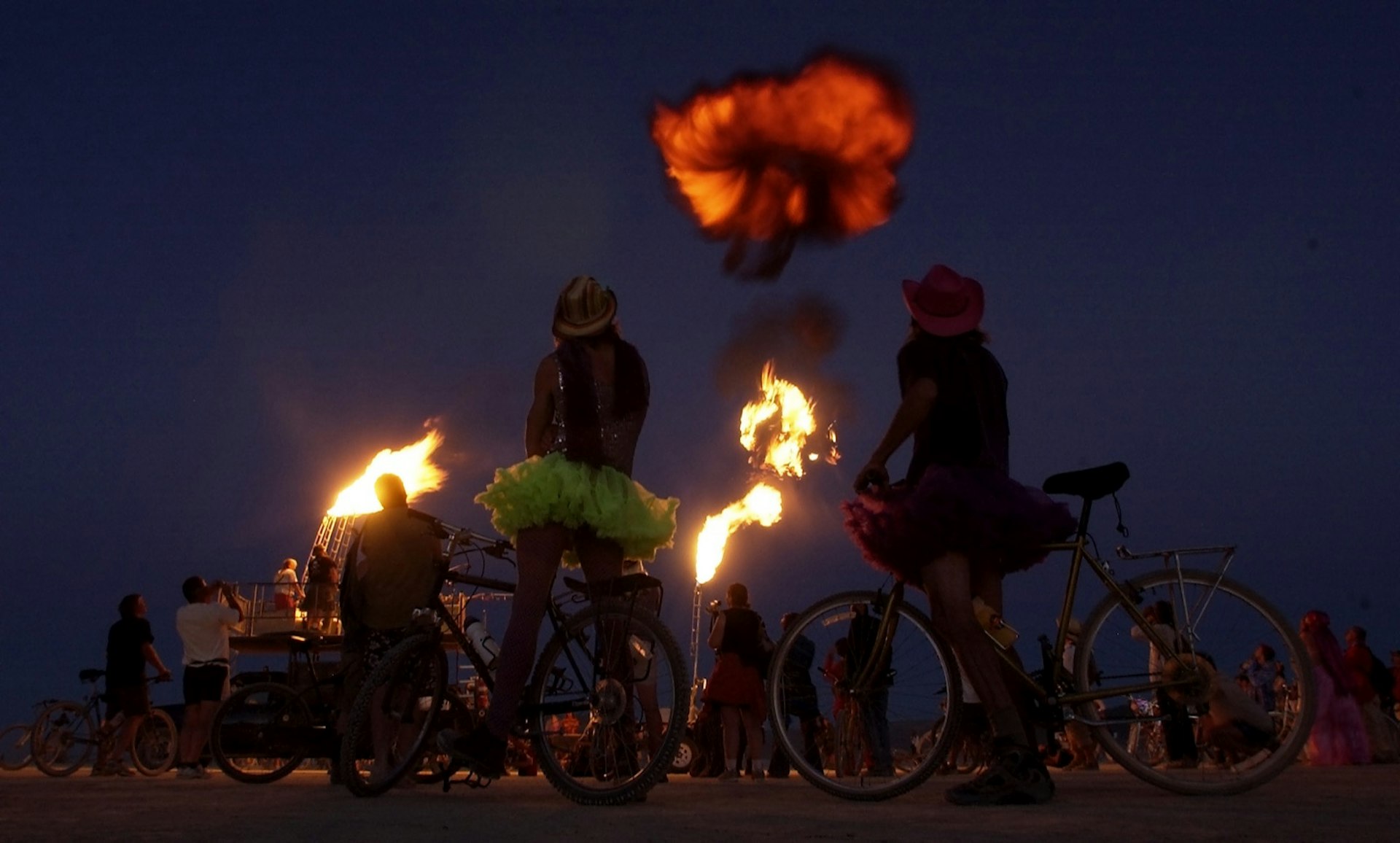 women in tutus watch a cloud of black smoke and flame rise into the desert night