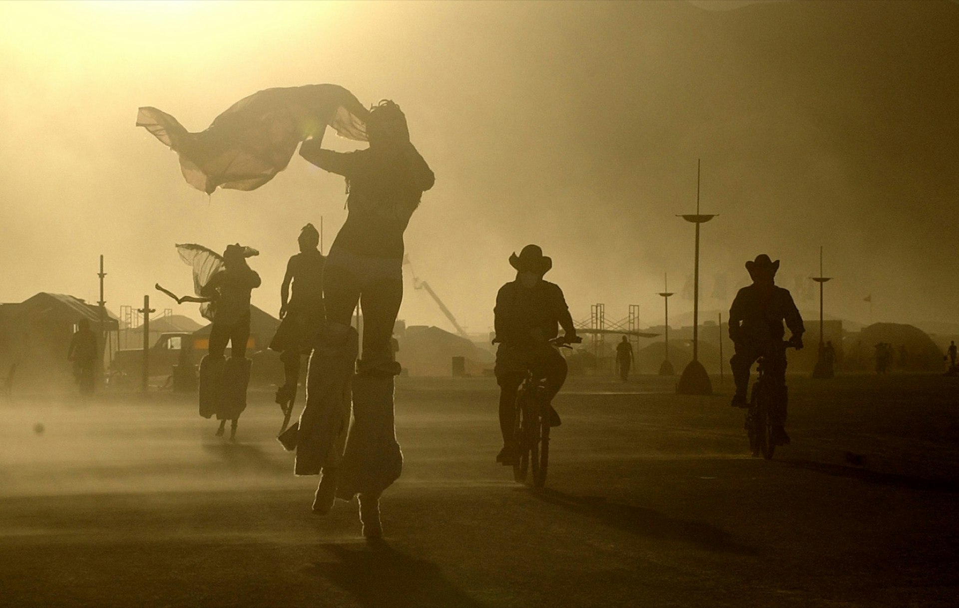 people walk on stilts and ride bikes in cowboy hats through a dusty desert day