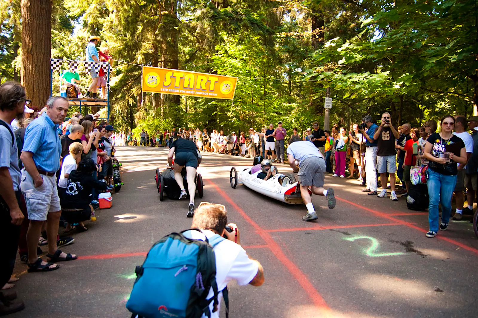 Features - Portland Soapbox derby race People push very sleek soapbox cars through a forested area on a road with lanes painted on it